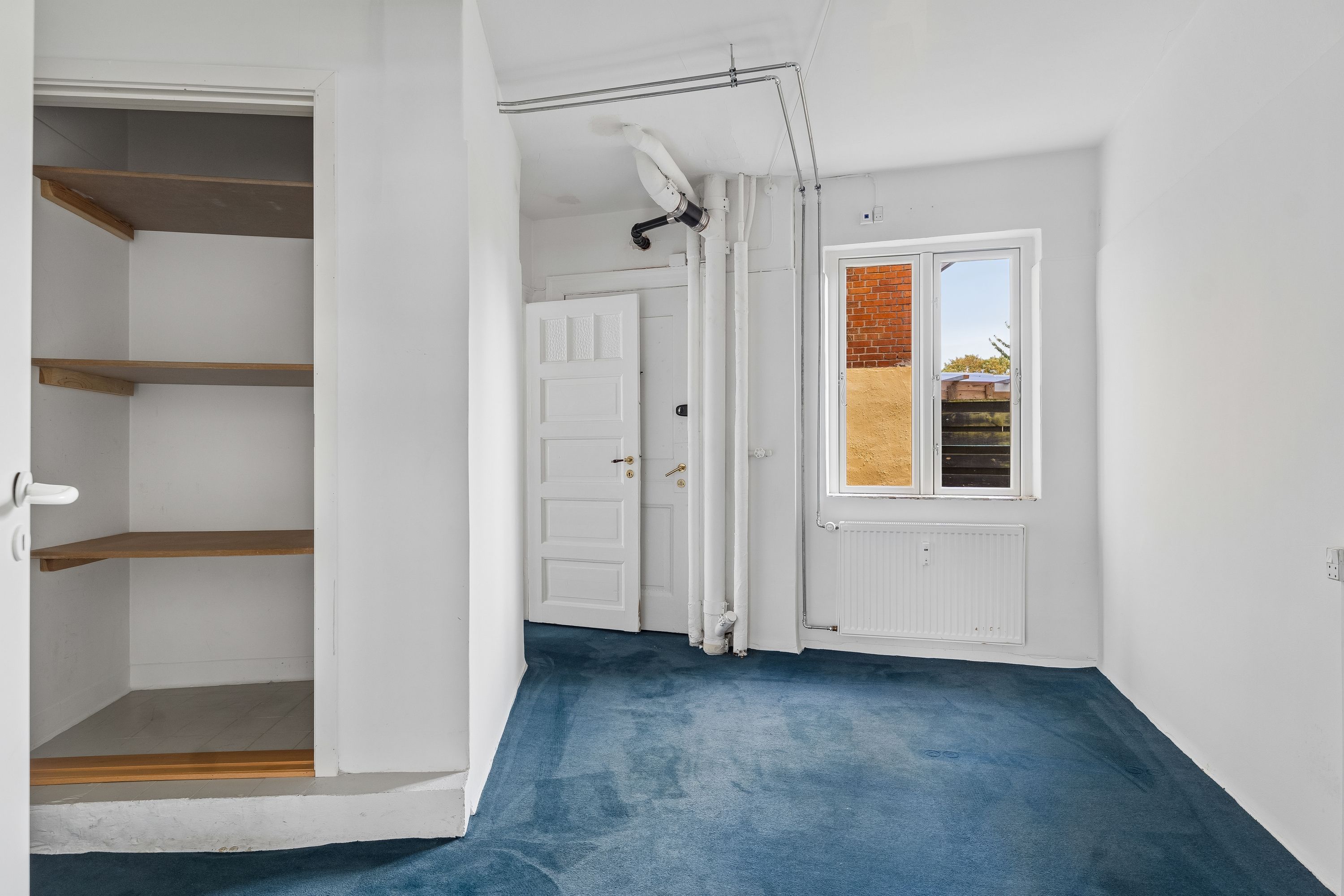 Kontorlokaler med hvidt vægpanel, lysegrå gulvbelægning, radiator og et vindue med udsigt til en mur. En hvid dør med guldfarvet håndtag og et rummeligt skab med tre træhylder