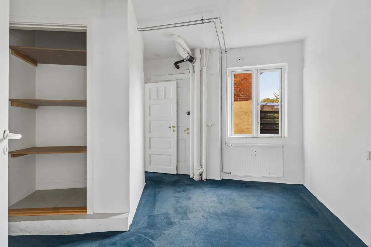 Kontorlokaler med hvidt vægpanel, lysegrå gulvbelægning, radiator og et vindue med udsigt til en mur. En hvid dør med guldfarvet håndtag og et rummeligt skab med tre træhylder