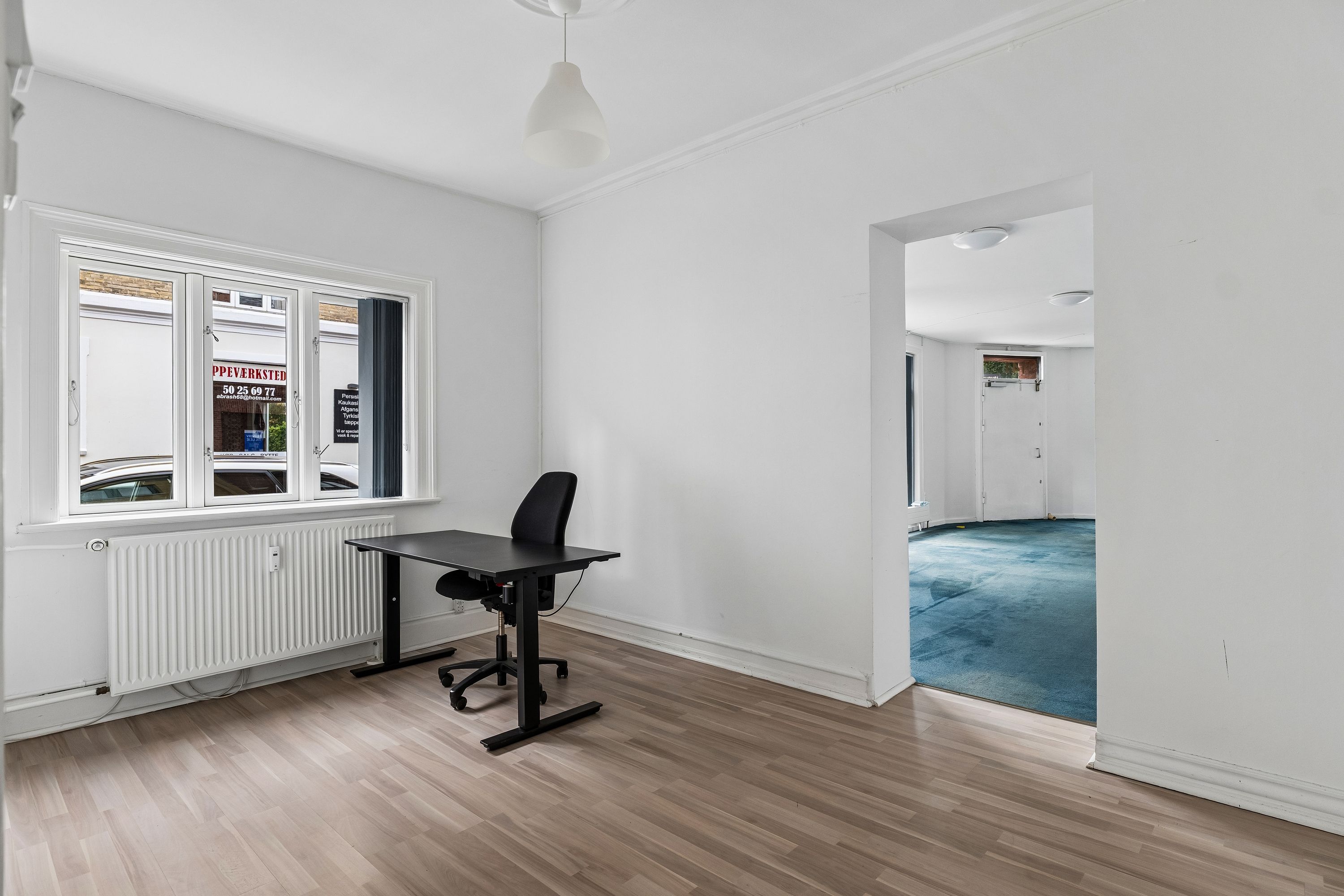 To store vinduer med glasdøre og et skrivebord med stol og hænglampe i en hvid stue med lysegrå gulvbelægning og hvide vægge