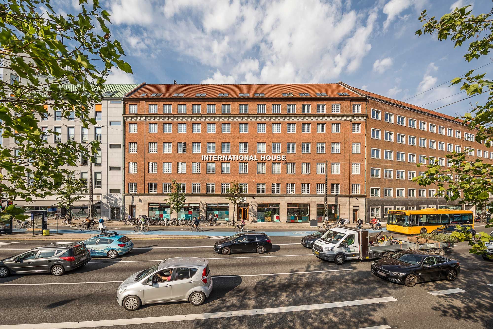 Foran International House kører biler og lastvogne, og en bus er parkeret. Her er offentlig transport, parkering og cykelstier.