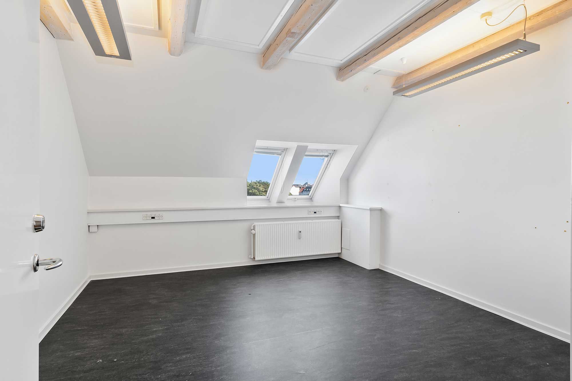 Store loftsrum med hvide vægge og lysegulv. Vinduerne er lukket, og der er et radiator og to lysekroner i loftet.