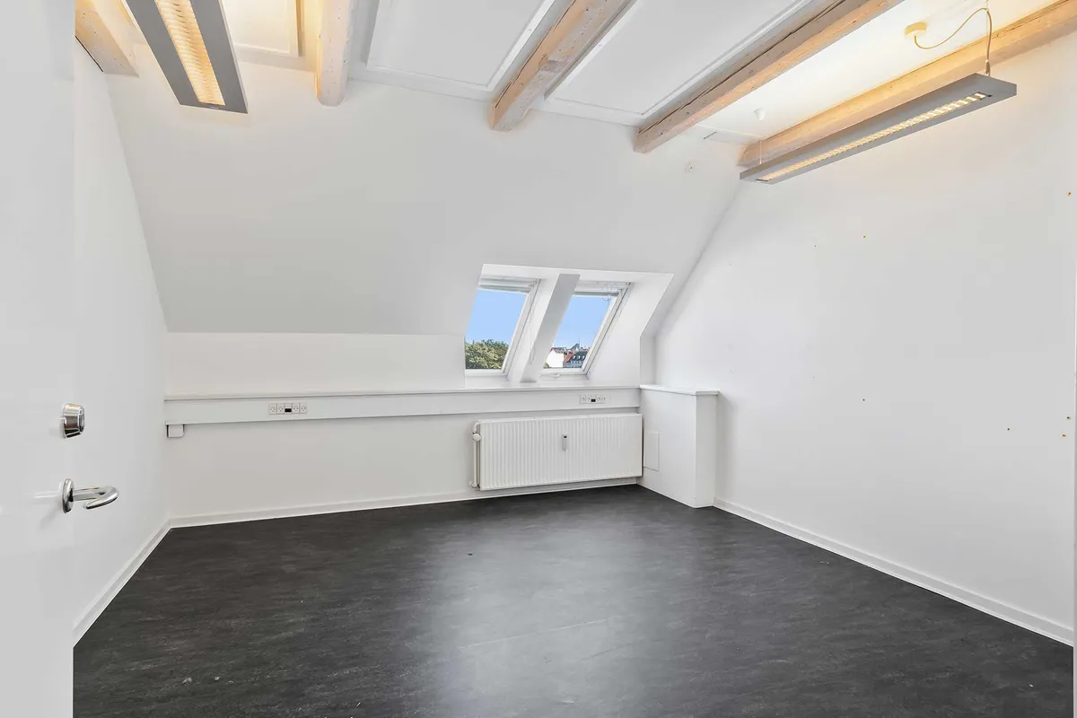 Store loftsrum med hvide vægge og lysegulv. Vinduerne er lukket, og der er et radiator og to lysekroner i loftet.
