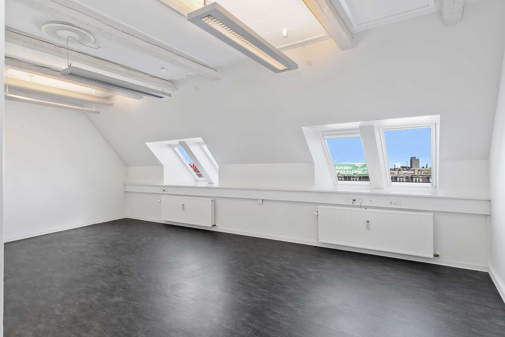 Køkken og lager i hjertet af København med moderne loftslejligheder, hvide vægge og moderne radiatorer samt tre store vinduer med udsigt til byens skyline.