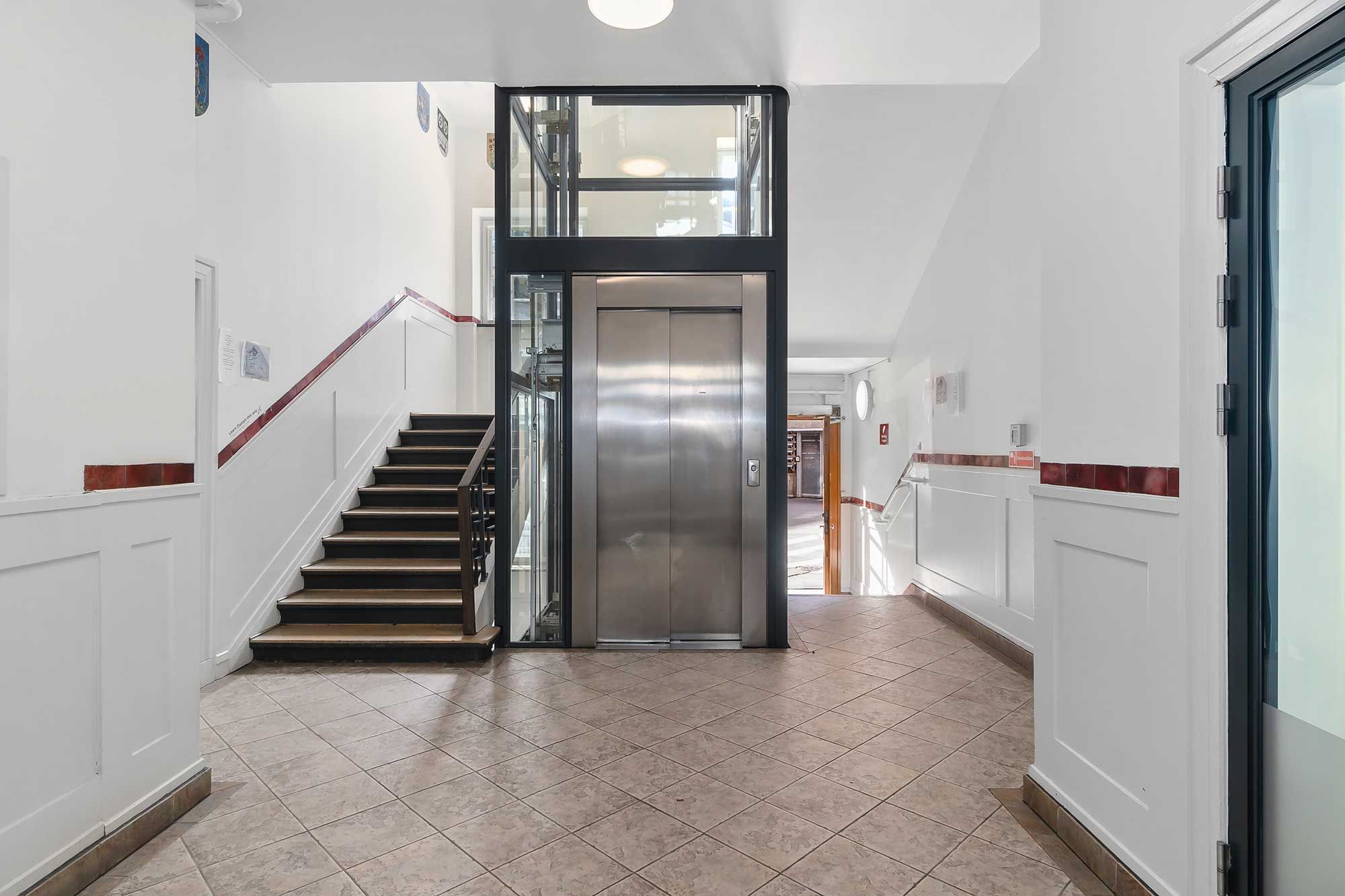 En indgang til et kontorhus med lysegule gulvbånd, hvidt panel og en elevator. Stigen viser gulvfarven, og der er skrift på væggen.