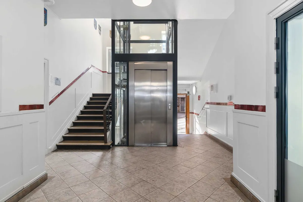 En indgang til et kontorhus med lysegule gulvbånd, hvidt panel og en elevator. Stigen viser gulvfarven, og der er skrift på væggen.