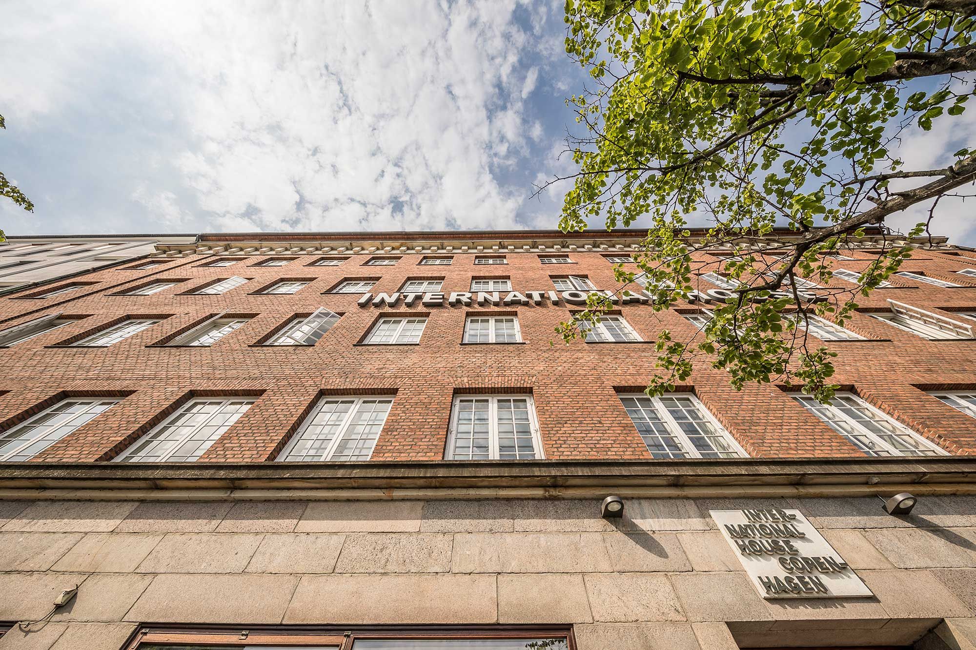 En stor, rød murstensejendom med store vinduer og en skjoldplakette med teksten International House. Over bygningen er der en stor, grøn trægren.