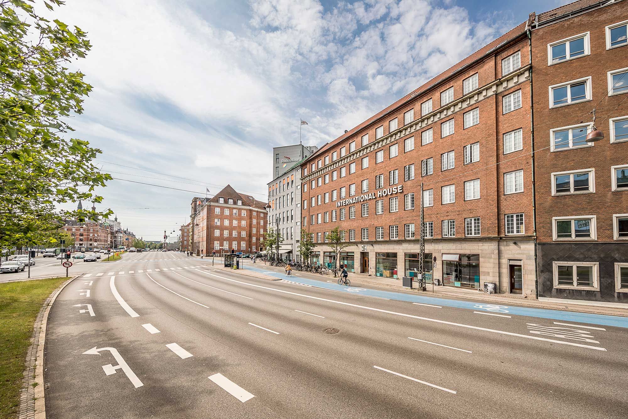 En stor, rød murstensbygning med International House-skilt står mellem grønne træer og en stor asfaltvej, som er let at komme til fra busser og biler