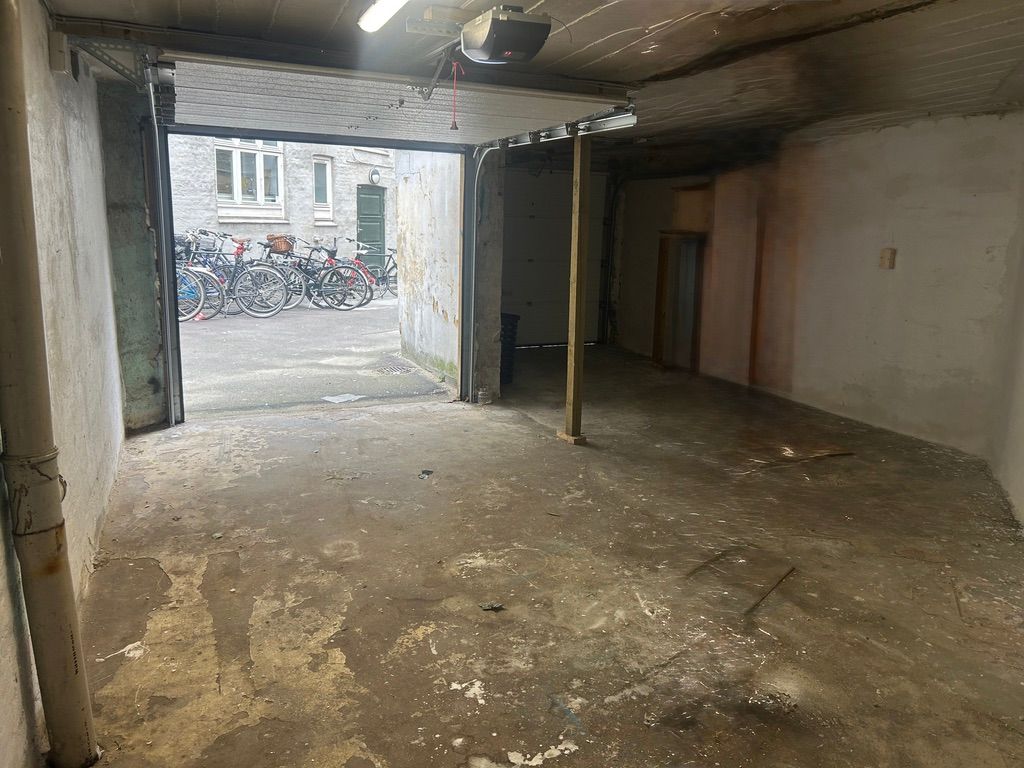 En gammel dobbeltgarage med åbne døre og flere cykler parkeret udenfor. Et højt lyskabin og en skrånende gulv med flere urene.