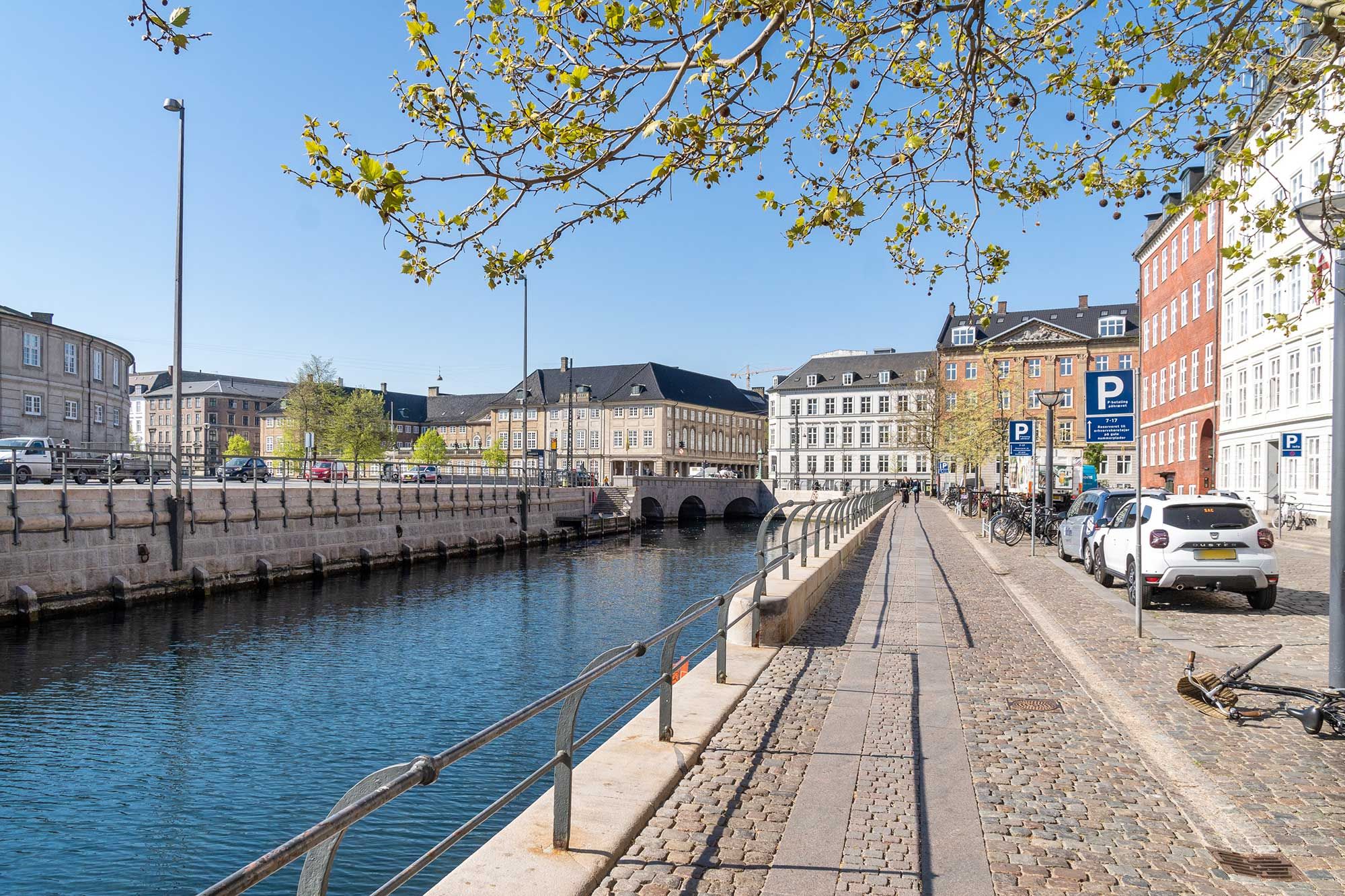 Charmerende baghus i Kompagnistræde, med brøndsdam og vandløb i baggården. Parkerede biler og cykler langs gaden, og smukke bygninger med parkerede biler og busser.