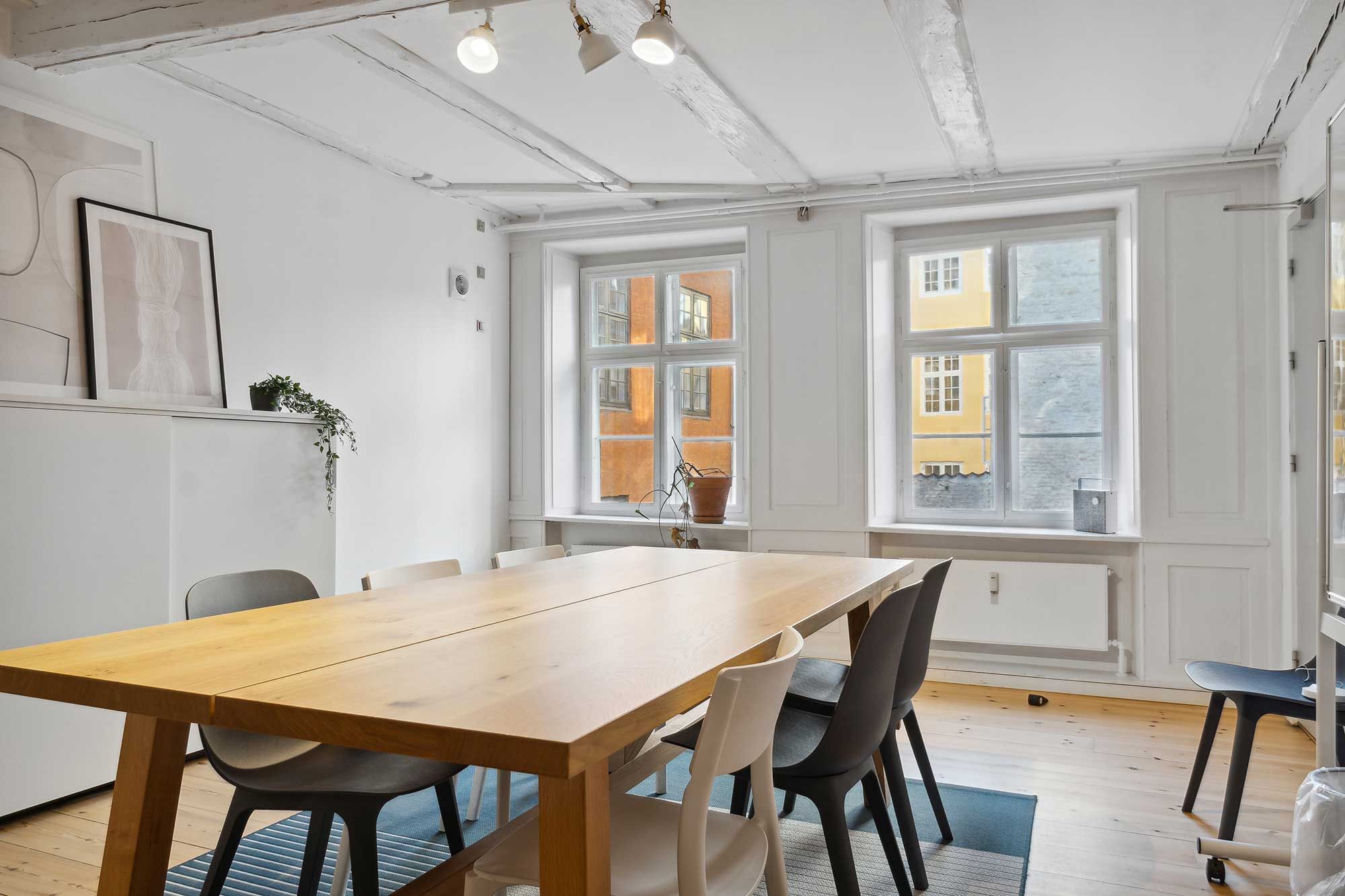 Kontorlokaler med lyse vinduer og lyse loftsbjælker. Massivt træbord med flere stole. Plante og kunstværk på væggen. Solrigt rum med trægulv.