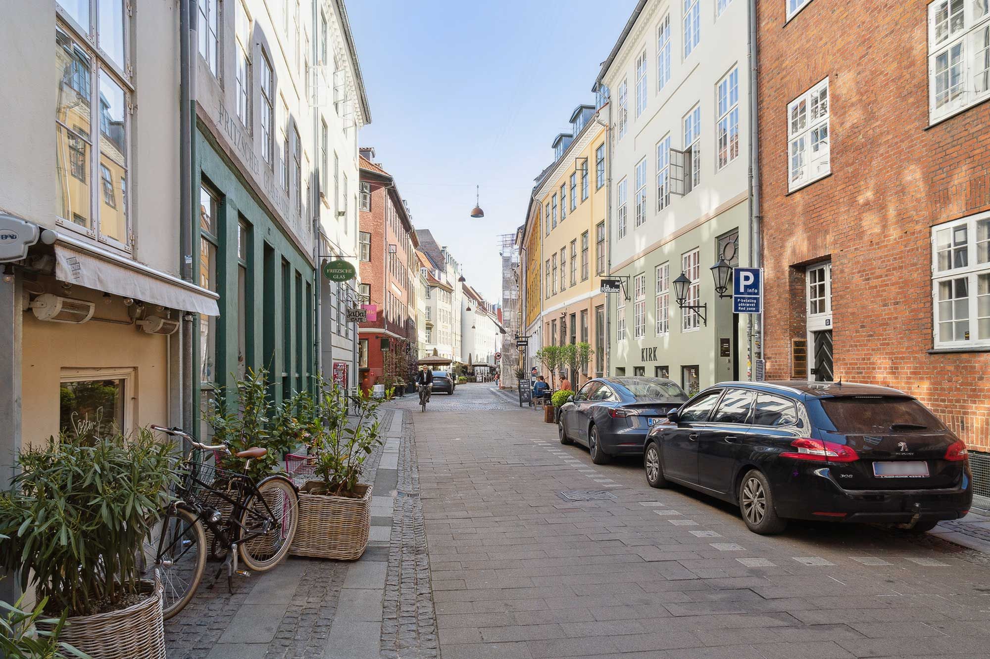 Kompagnistræde, København med to parkerede biler, cykel, buske, vinduer og facader med røde mursten og grønne ruder.