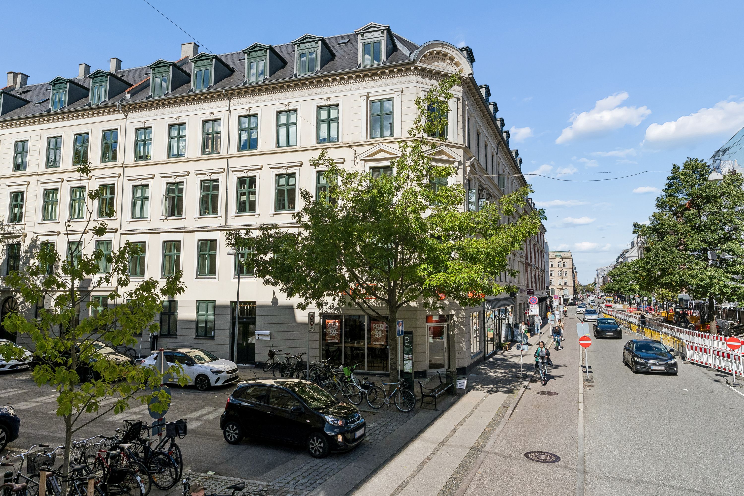 Moderne bygning med høj loftshøjde og mange vinduespartier på Roarsvej i Frederiksberg. På gågaden er der en cykelskur med parkerede biler og gående folk