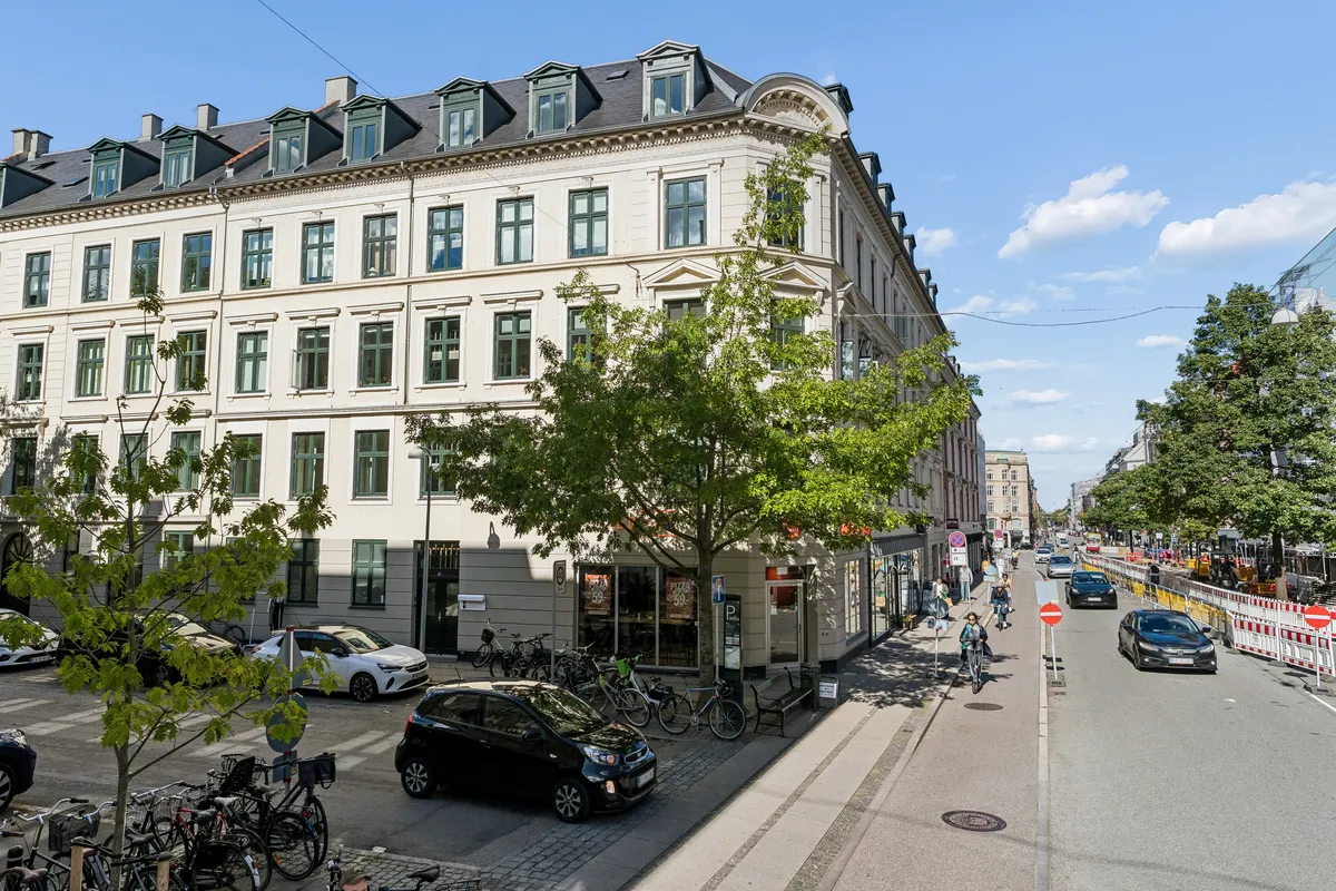 Moderne bygning med høj loftshøjde og mange vinduespartier på Roarsvej i Frederiksberg. På gågaden er der en cykelskur med parkerede biler og gående folk