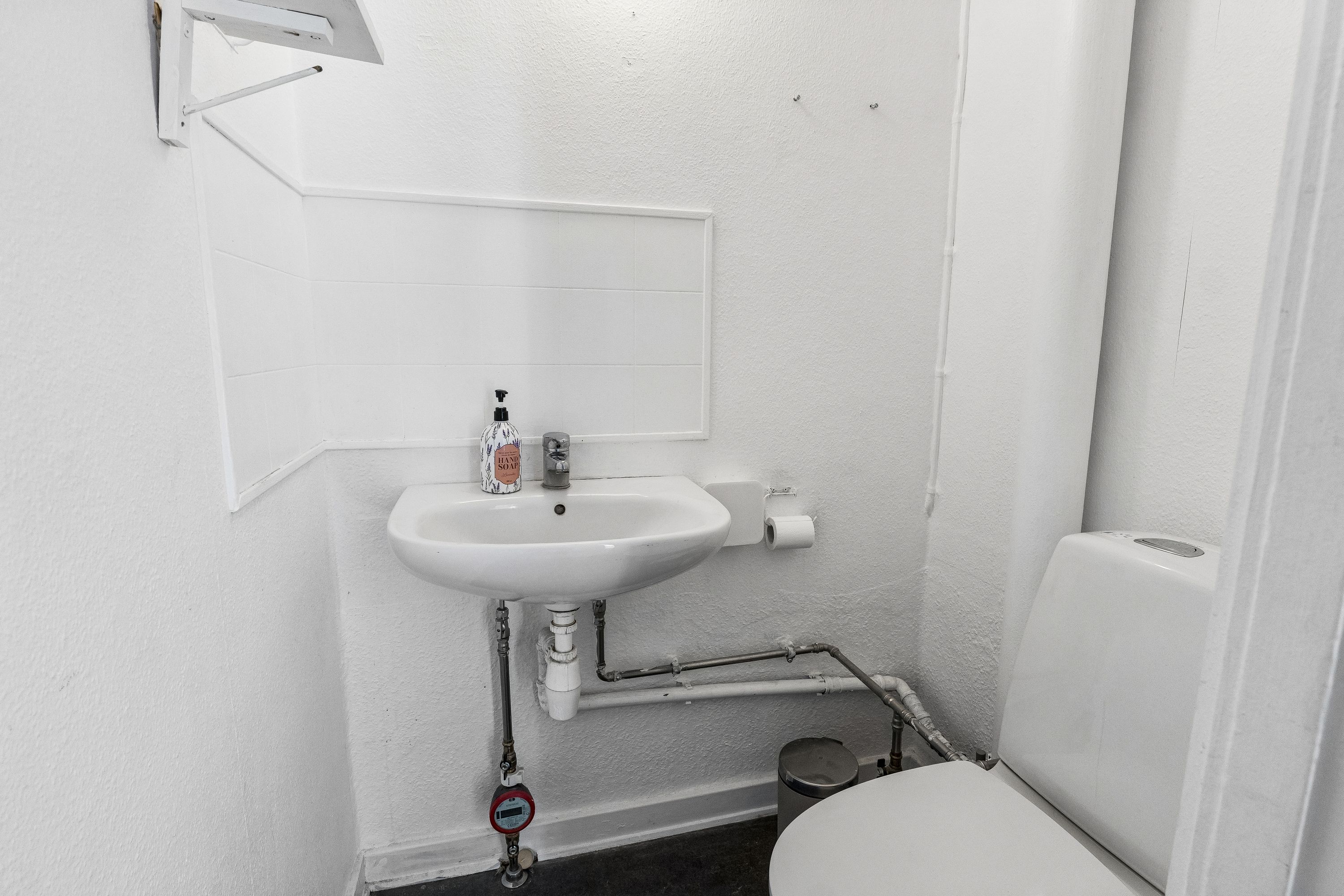 Toalett med hængetoilet og hvid, rektangulære vask med håndsæbeflaske og håndklæderol. Toilet med gulvmonteret, rundt affaldskurv og en redningsventil.
