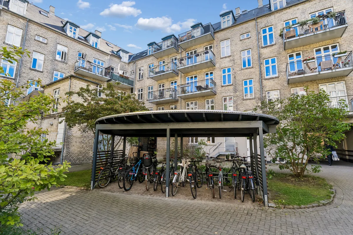 En parkeringsplads med flere cykler under et metaltage. En stor, brun bygning med flere balkoner og vinduerne i række. En person i baggrunden og nogle grønne planter foran.