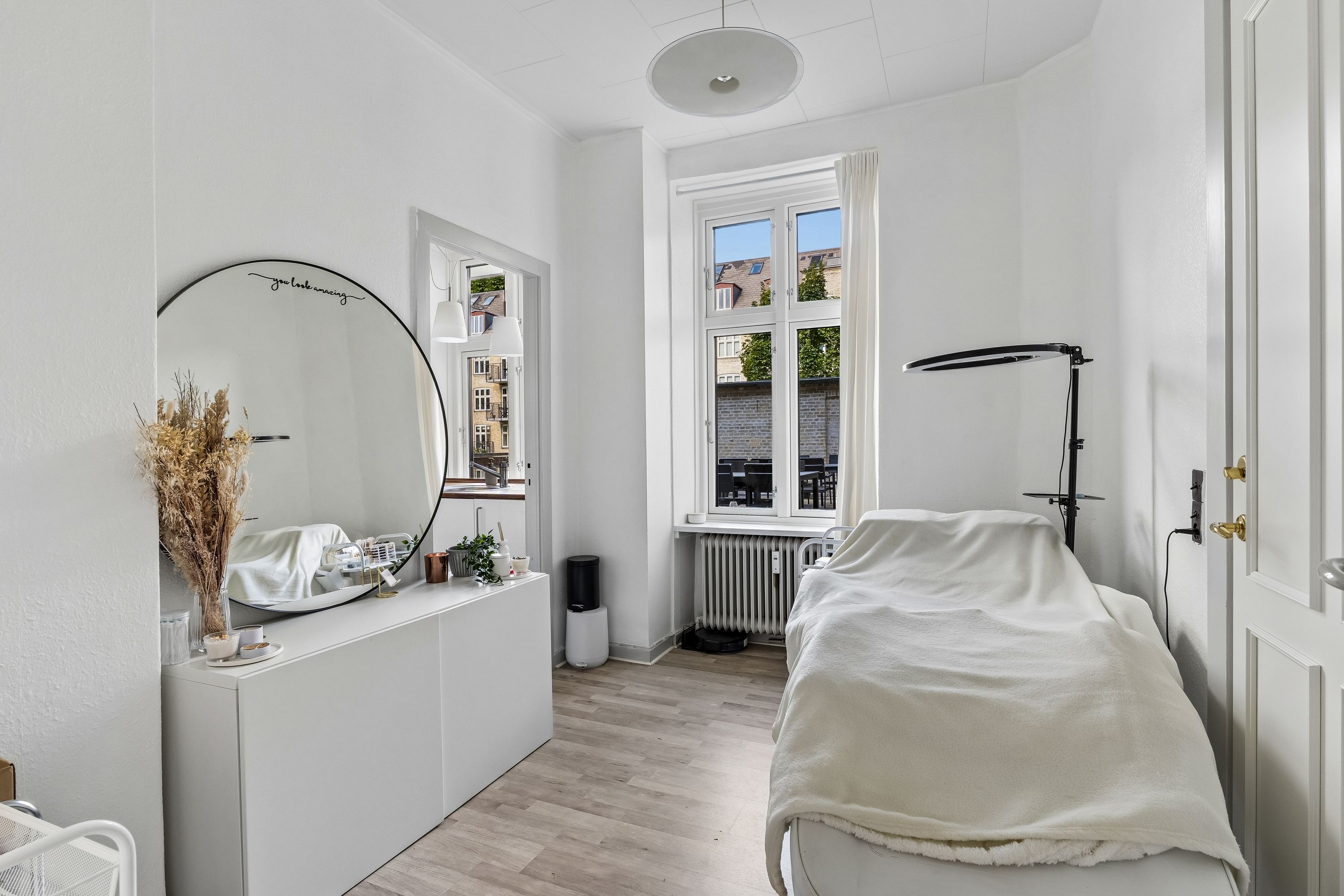 Luksuriøs kontor med høj loftshøjde, vinduer med udsigt til grønne områder, en stor rundspejl, et hvidt bord med dekorationer, et højt stativ og en lampe