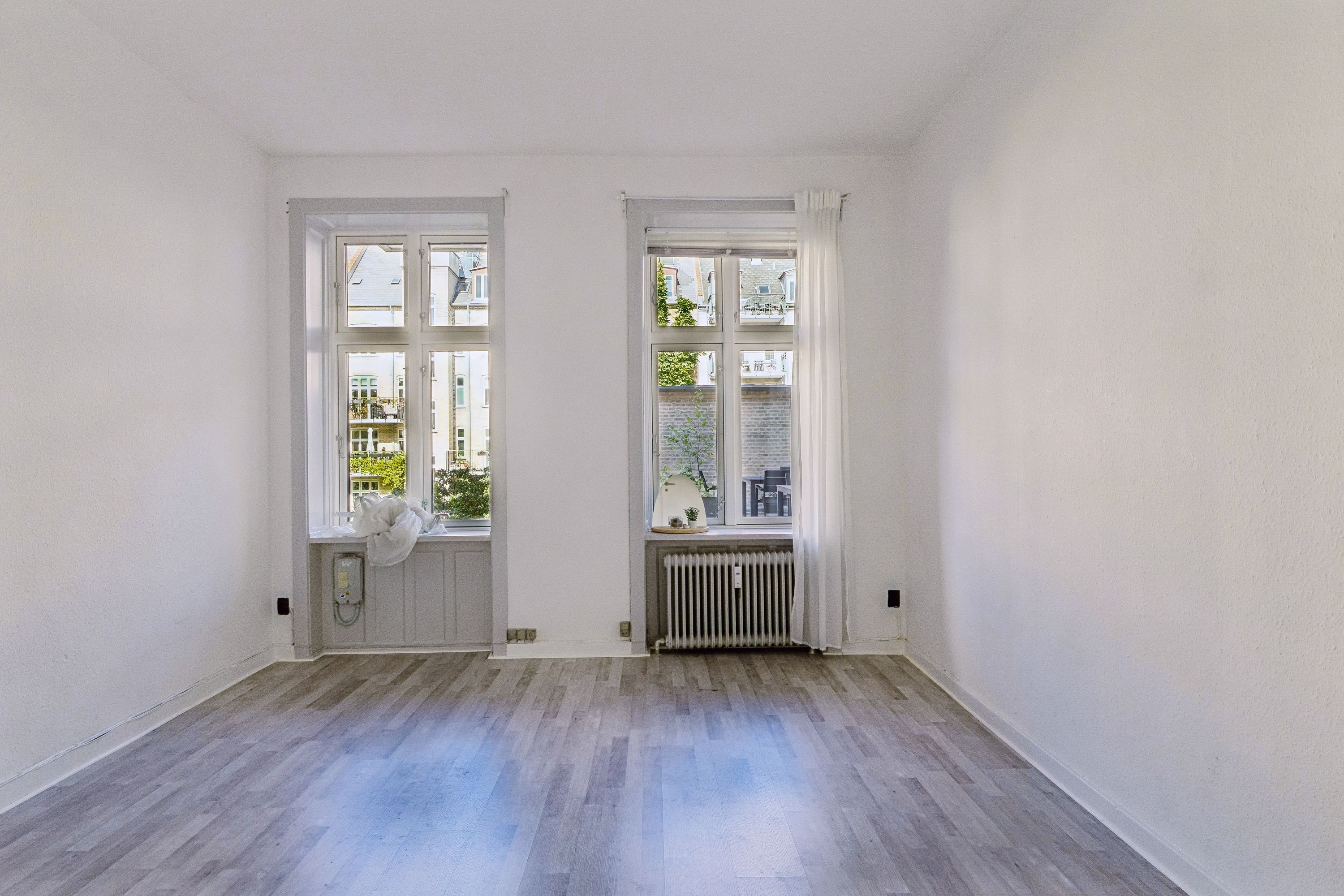 Et tomt kontor med højloft og lyse vinduespartier til leje på Roarsvej i Frederiksberg. Væggene er hvide, gulvet er træ og der er radiatorer installeret under vinduerne