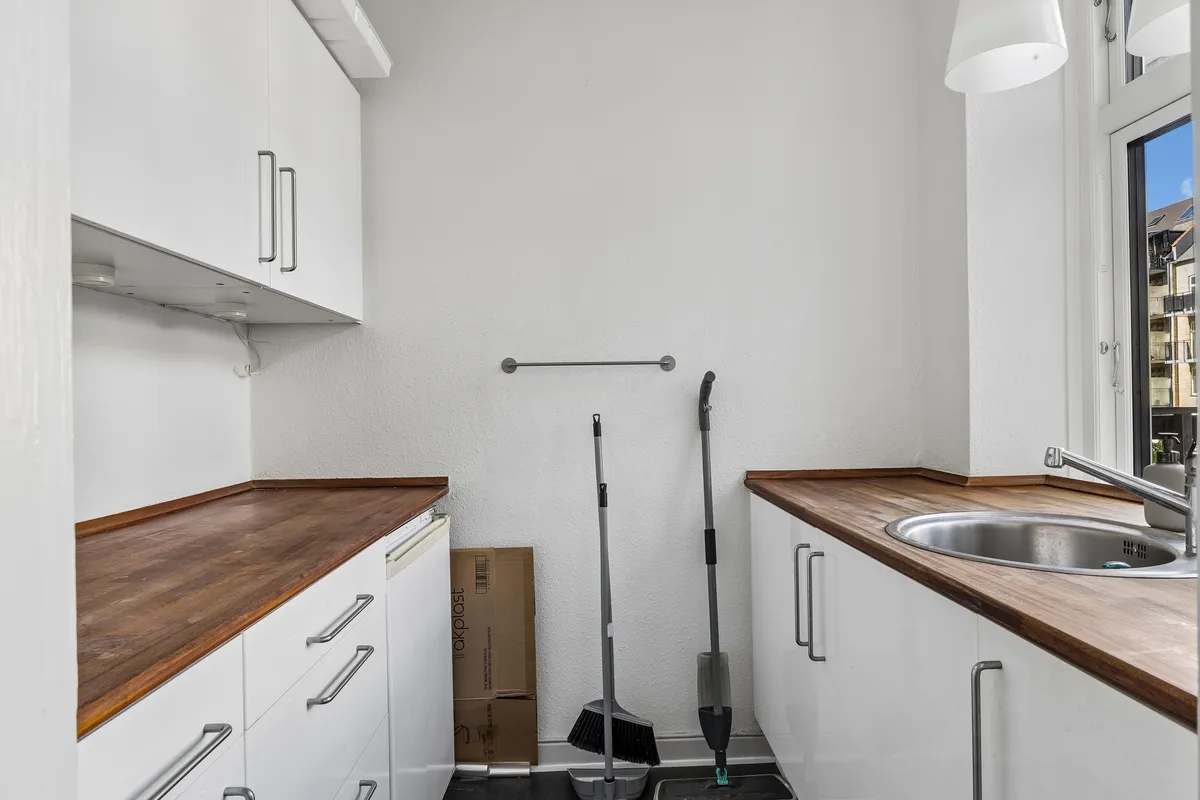 Ren, hvid vaskerum med moderne, lyse trægulve og møbler. Rent og funktionelt med vask, spidse tørrestang og rengøringsudstyr. Vindue ud mod lys og frederiksberg-bygninger.