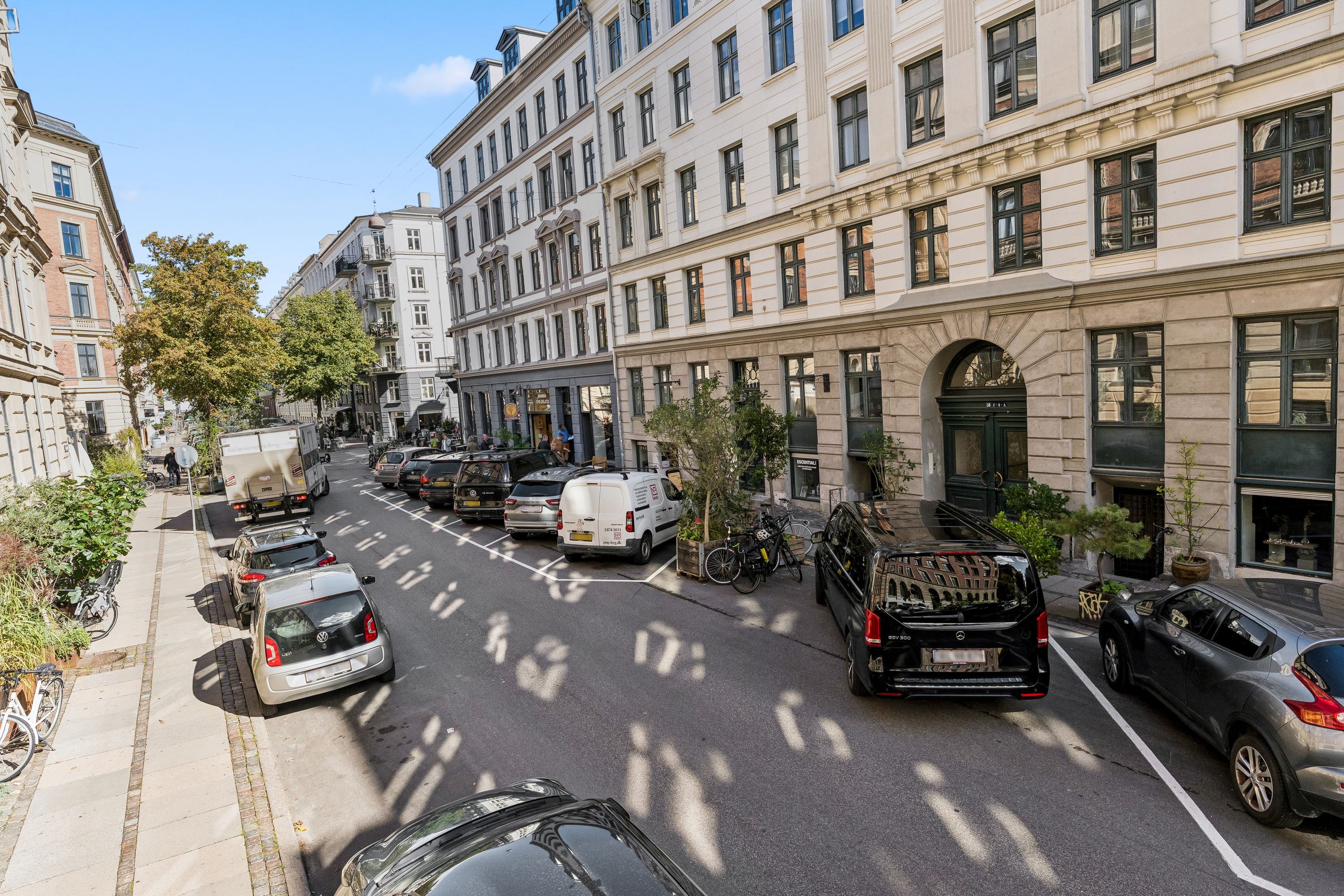 Københavnsgade med biler og cykler parkeret og en bygning med flere vinduer og en dør med en høj plante foran