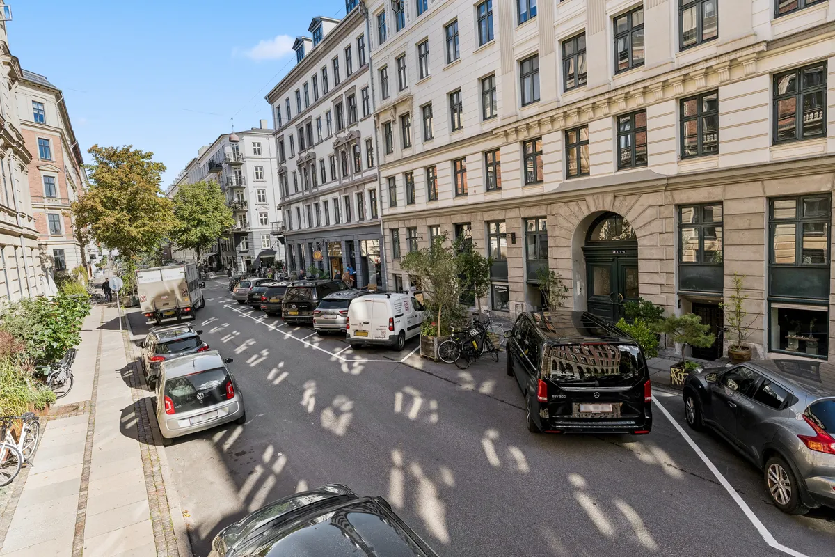 Københavnsgade med biler og cykler parkeret og en bygning med flere vinduer og en dør med en høj plante foran
