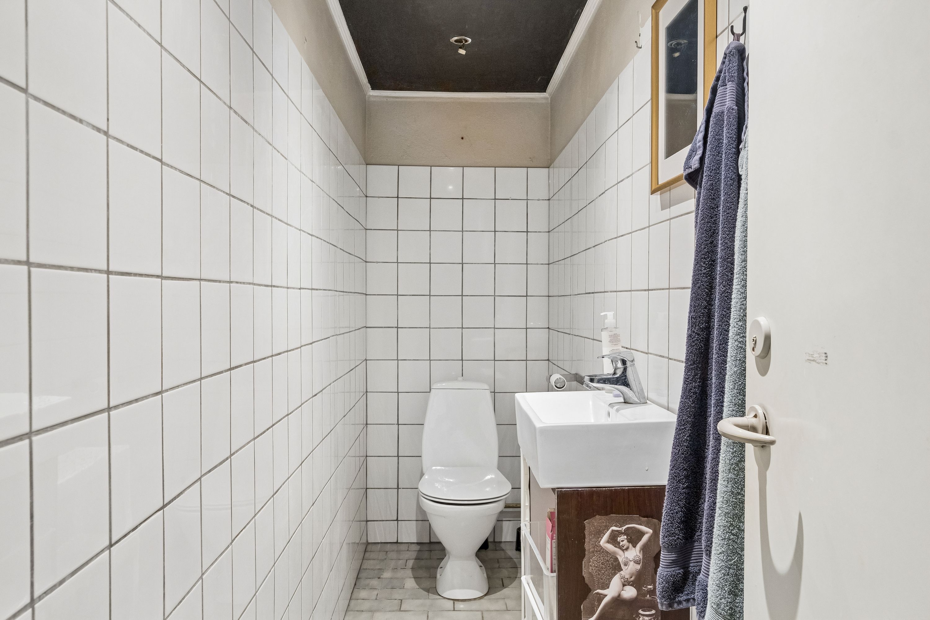 En lille, hvidt flisede badeværelse med toilet, håndvask og tørklæder samt søjle på loftet, der har et lysekron