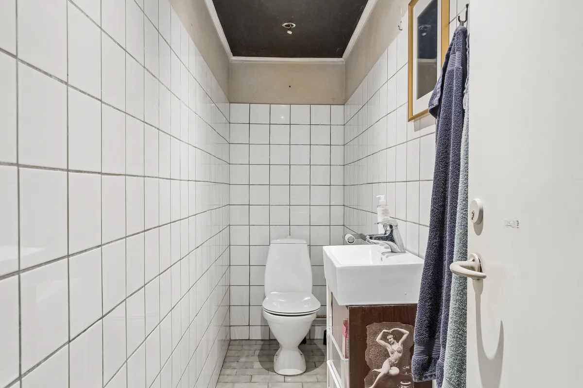 En lille, hvidt flisede badeværelse med toilet, håndvask og tørklæder samt søjle på loftet, der har et lysekron