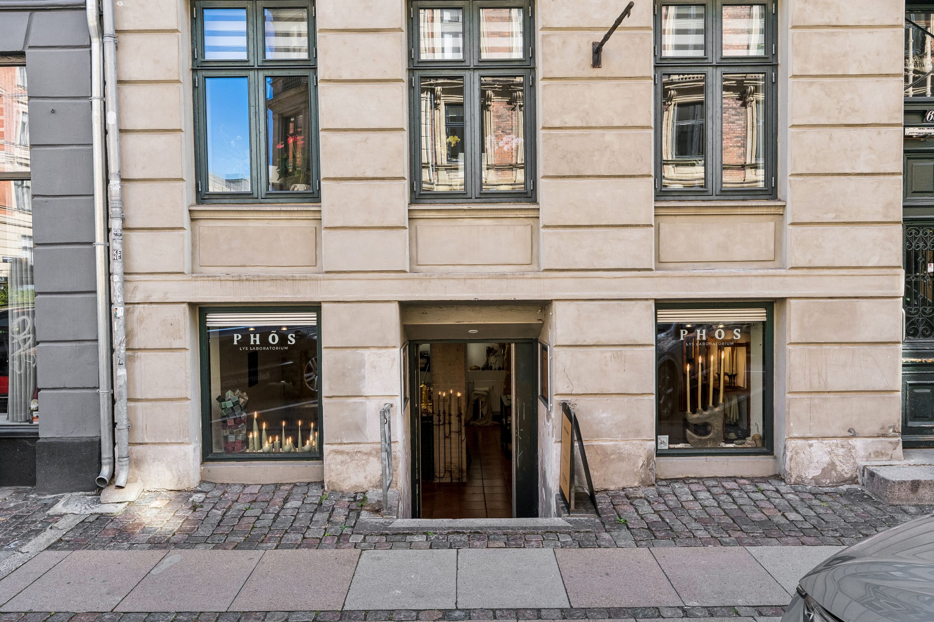 Kælderleje på Nansensgade 60 i København K. Lejlighed med moderne interiør og tre vinduer. Indkøbsmuligheder og butiksvinduer i forgrunden. Fortov med parkerede biler og fortovsfortov.