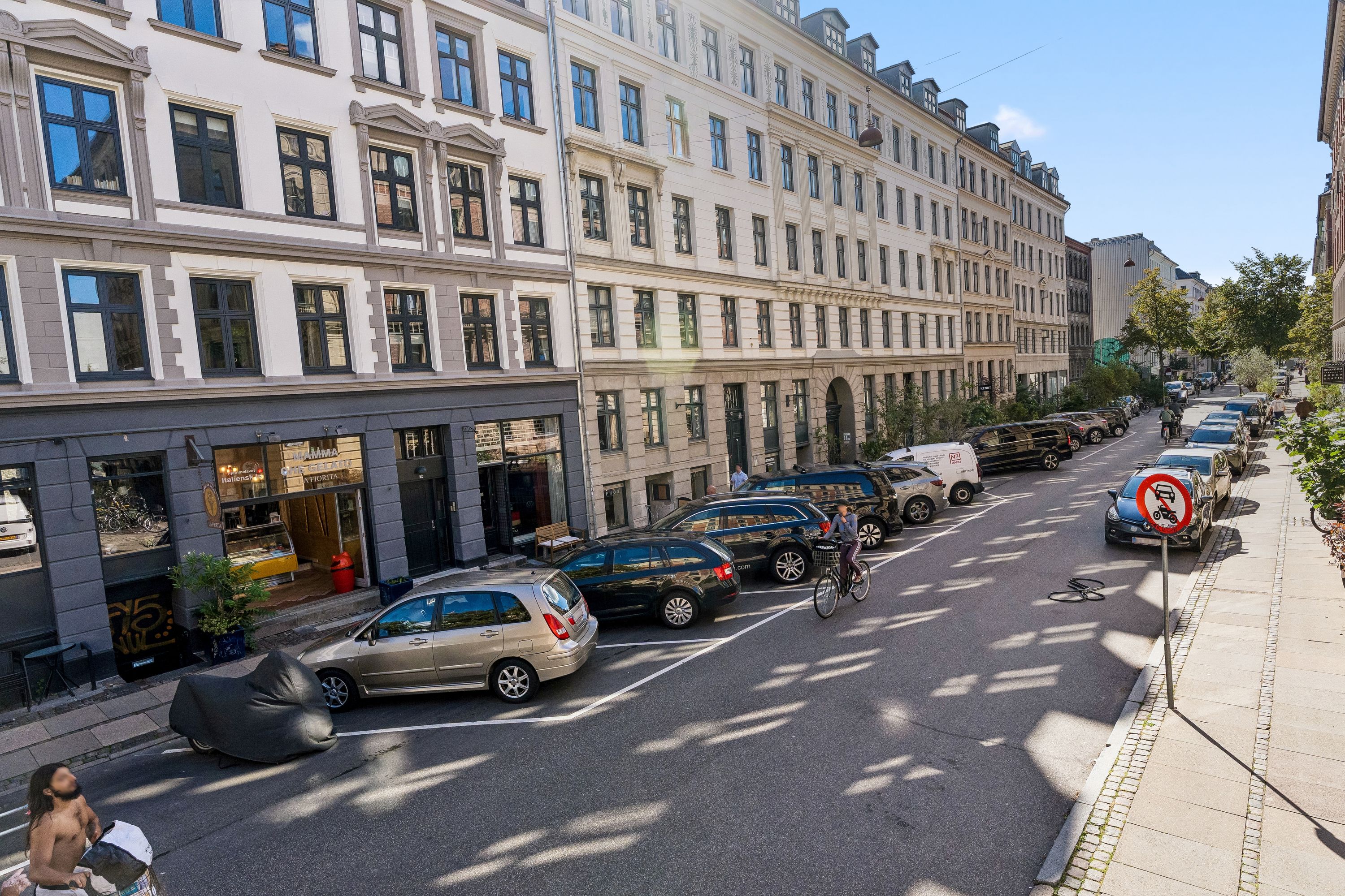 En københavnsk gade med parkerede biler, cykelstier, gamle huse og grønne planter i baggrunden og en mand med en pose