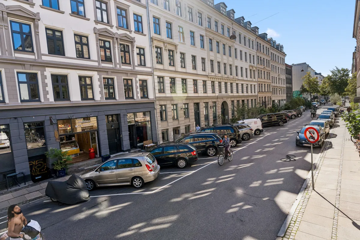 En københavnsk gade med parkerede biler, cykelstier, gamle huse og grønne planter i baggrunden og en mand med en pose