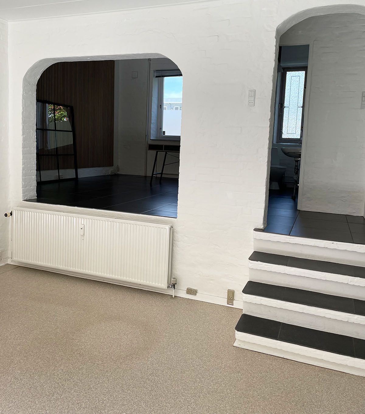 Rummelig lejlighed med to vinduer, hvid væg og mørke gulv og hvid radiator