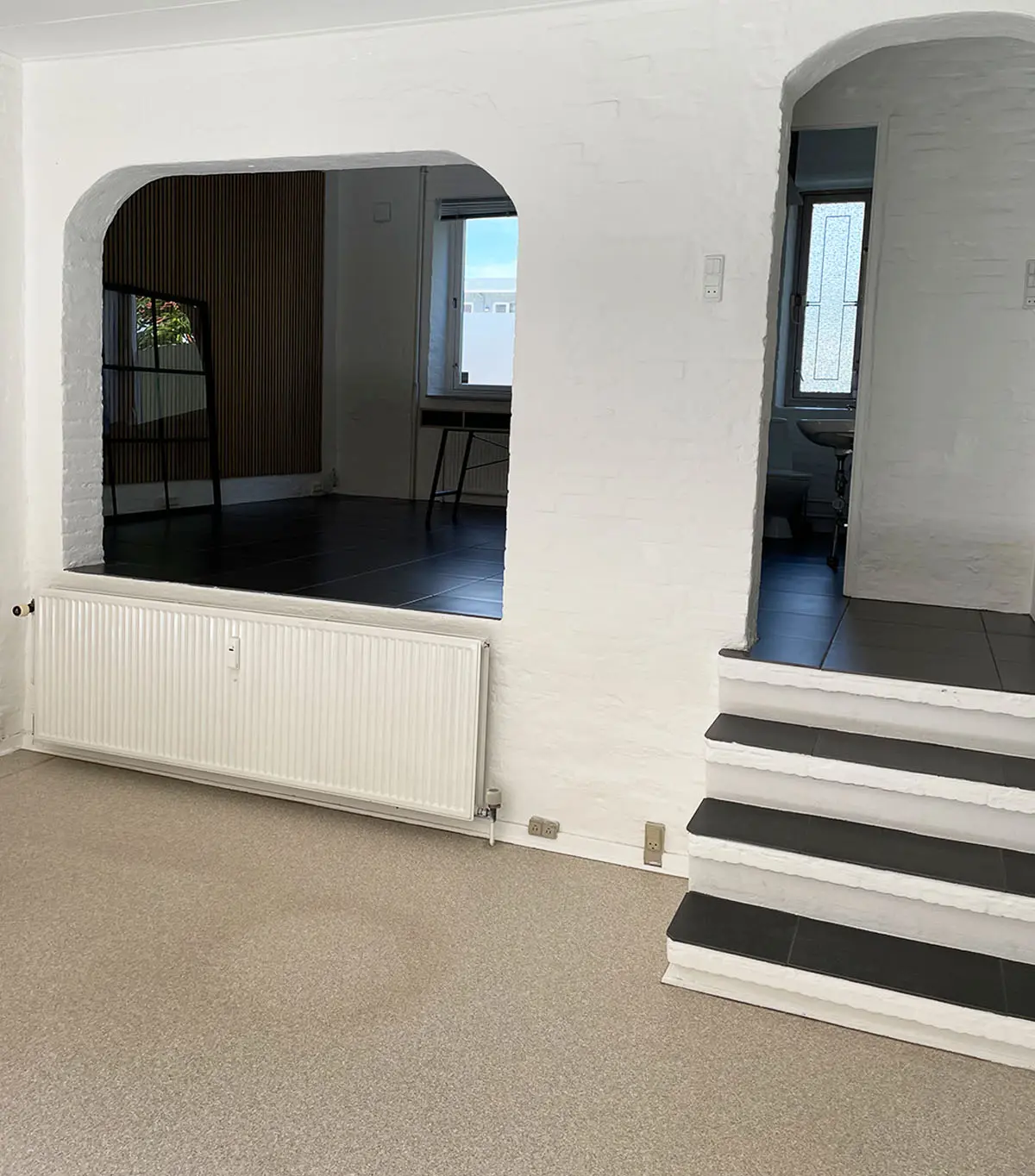 Rummelig lejlighed med to vinduer, hvid væg og mørke gulv og hvid radiator
