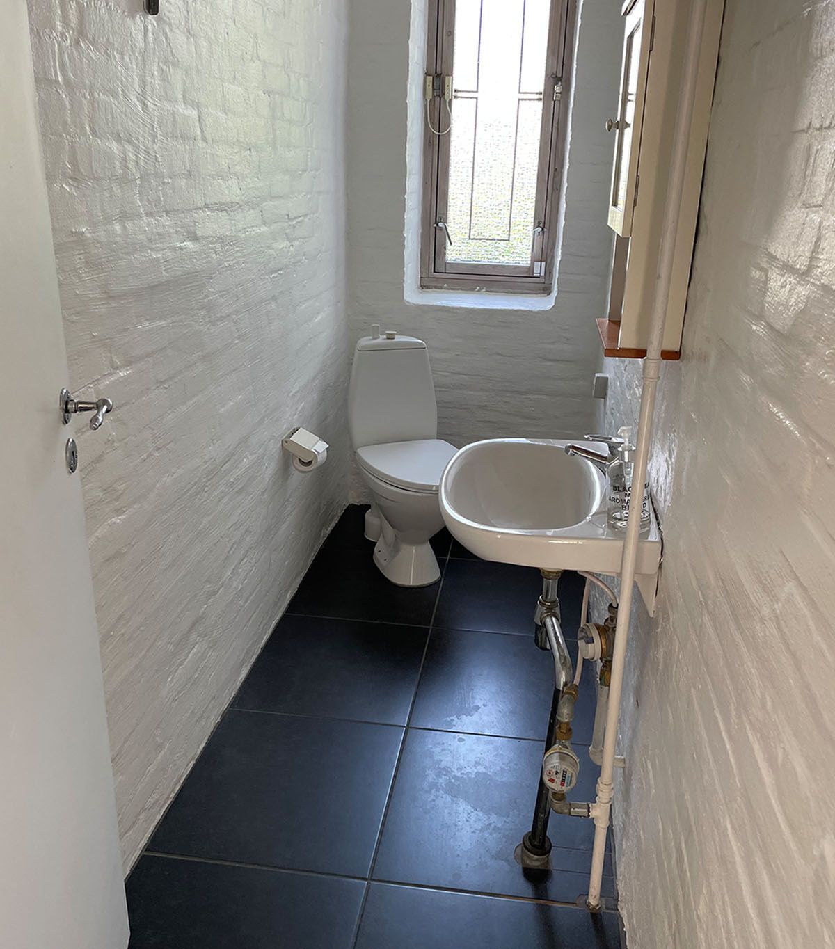 Et lille hvidt badeværelse med toilet, håndvask og et vindue med udsigt til gaden