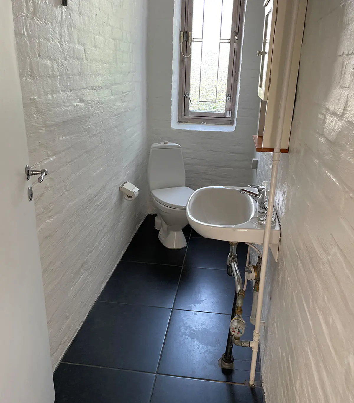 Et lille hvidt badeværelse med toilet, håndvask og et vindue med udsigt til gaden