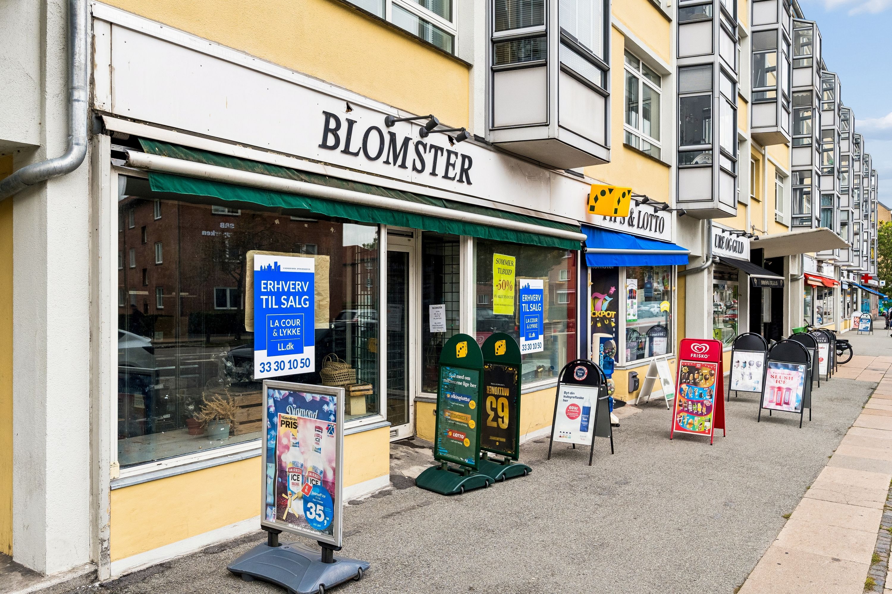 Frederikssundsvej 306B i Brønshøj med Blomster-butik og adskillige reklameskilte og butikker. Lysene er tændt, og der er mange vinduer med reklamer og annoncer.
