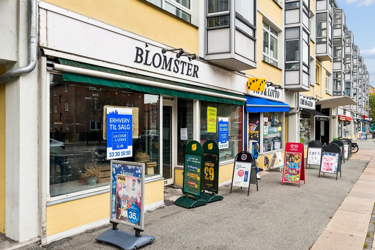 Frederikssundsvej 306B i Brønshøj med Blomster-butik og adskillige reklameskilte og butikker. Lysene er tændt, og der er mange vinduer med reklamer og annoncer.