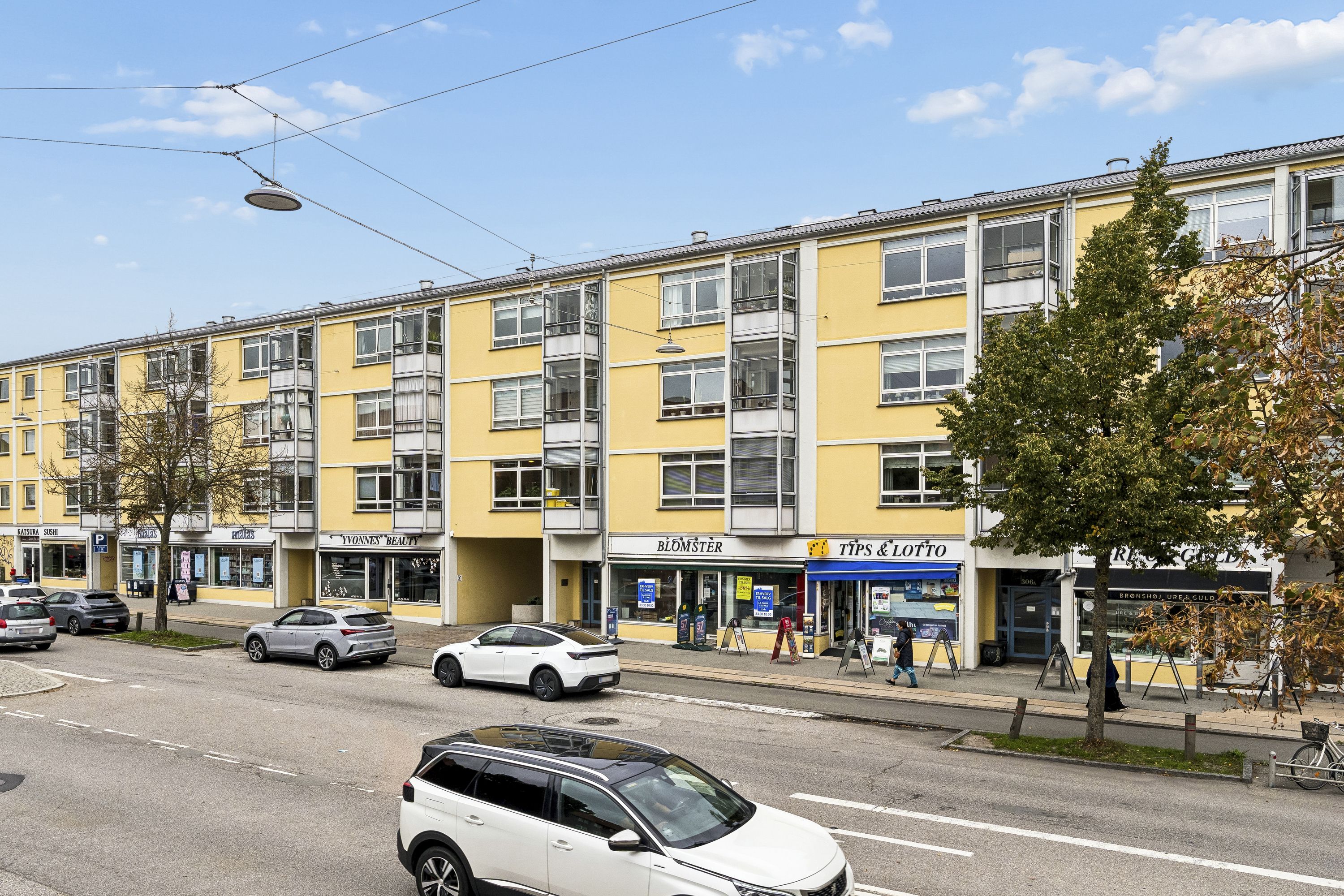 To biler kører på Frederikssundsvej foran et stort gule lejlighedsbyggeri med butikker og kontorer. Mennesker går forbi, og en cykel står på fortovet.