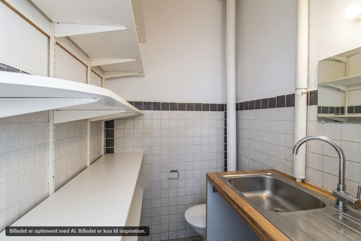 Et lille og rent badeværelse med en hvid toilet, håndvask og spejl. Der er hvide hylder og hvidt gulv. Der er en hvid toiletpapirholder og en køkkenskraldsspand.