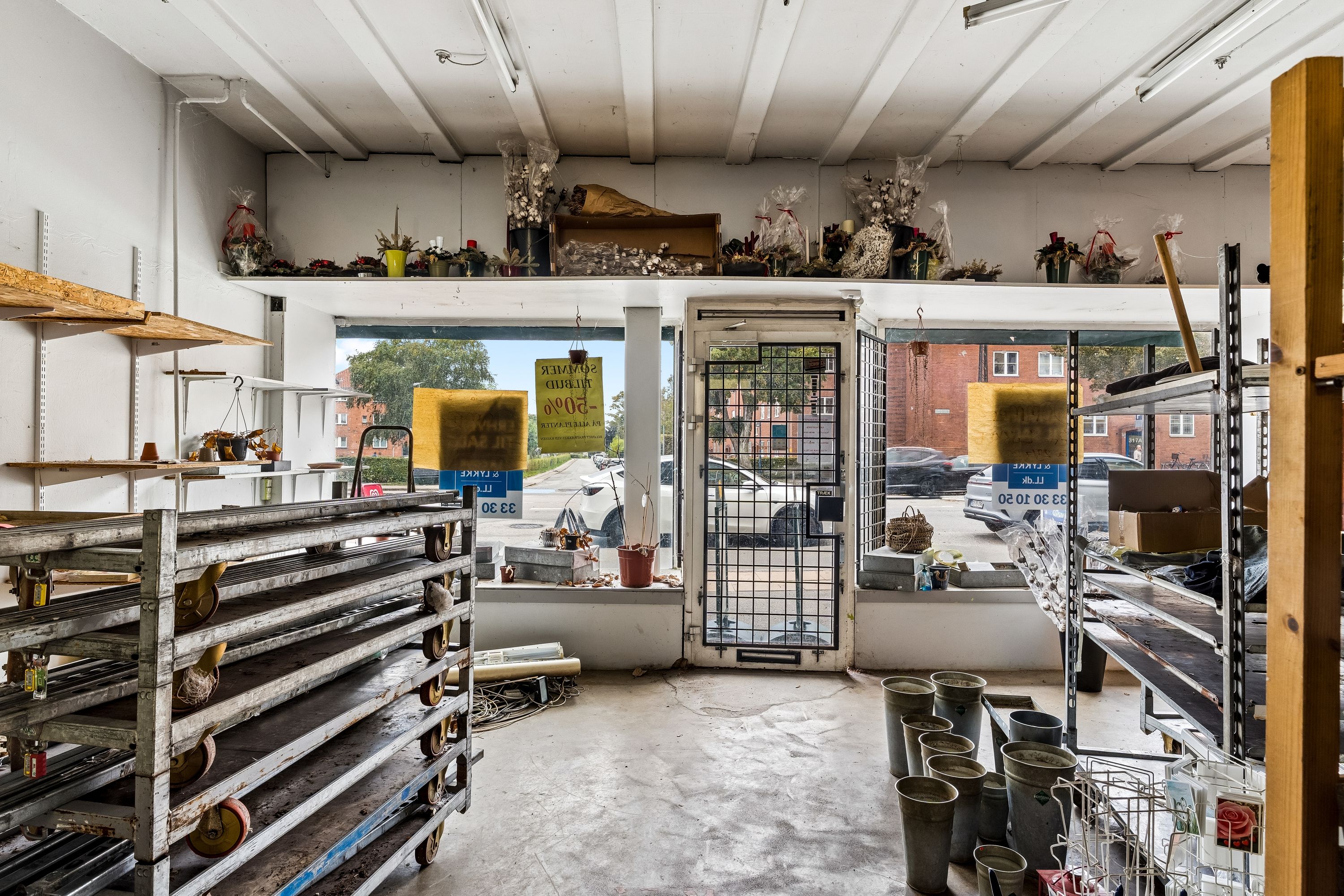 Forretningslokal med smykke og blomster på hylder og vinduer. Rækkevis af smykker og blomster i potter og beholdere i forretningen. Kasser og kurve med produkter i forretningen. Parkerede biler og bygninger på gaden udenfor.