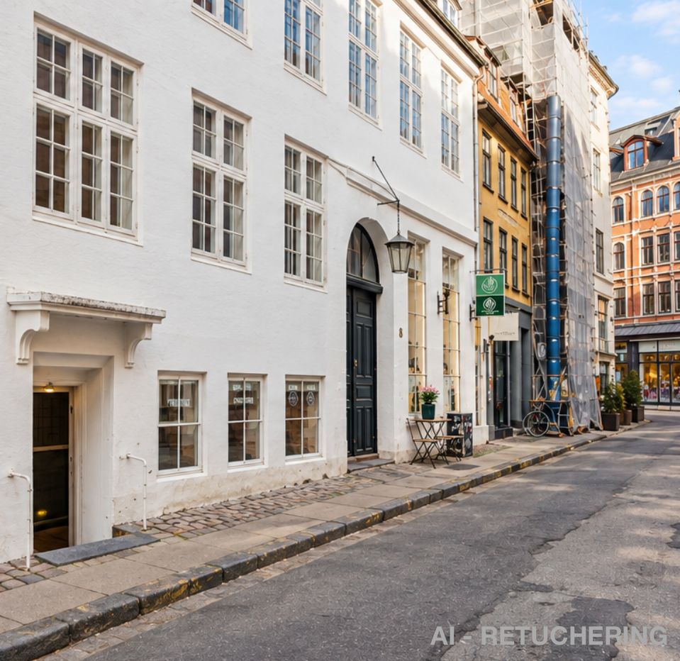 En ældre bygning med hvide vægge og flere vinduer står på Badstuestræde i København. Der er et bord med blomster på siden af gaden og en cykel parkeret ved en bygning