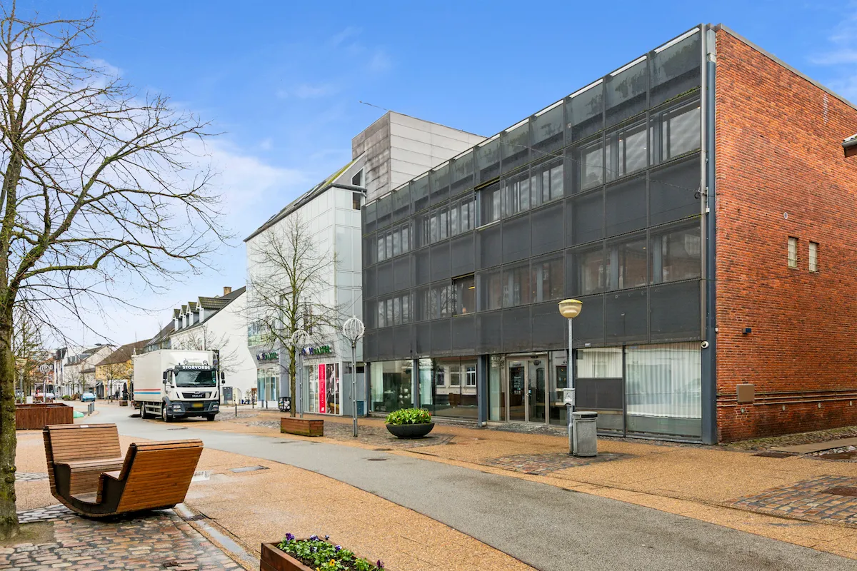 En hjørnebygning med røde mursten og glasvinduer i Hjørring. En stor lastbil kører langs gaden. Der er træbænke og grønne plantager uden for bygningen.