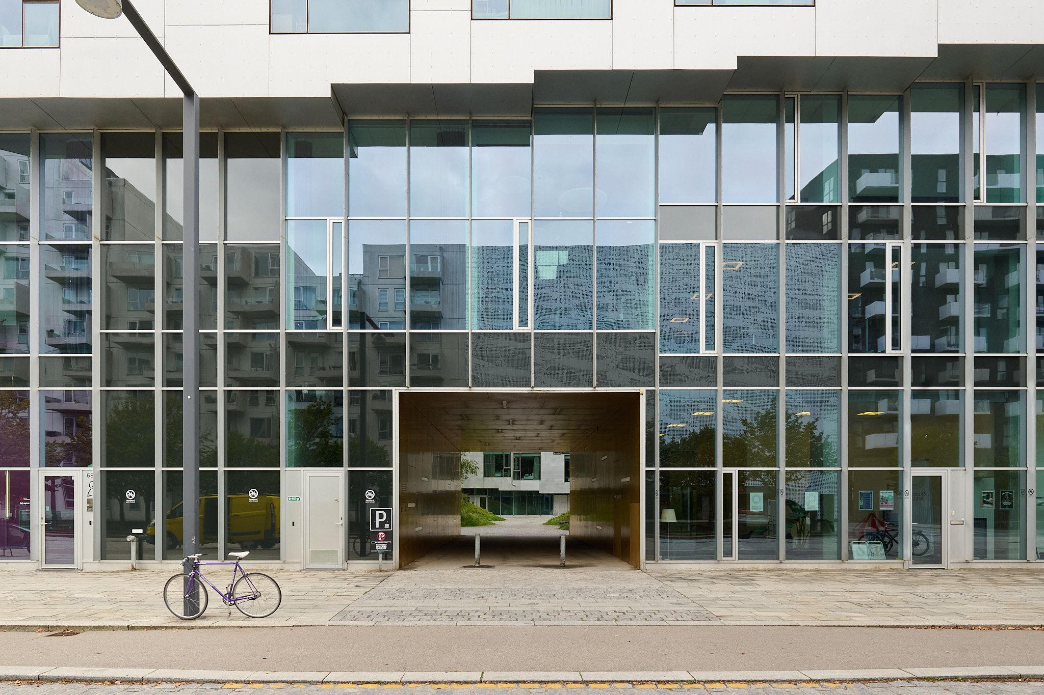 Moderne glasbygning med stor garageindkørsel og parkeringsplads foran. Glasvinduer, stålsøjler og parkerede cykler i forgrunden. Tidligere kontorlokaler i baggrunden.