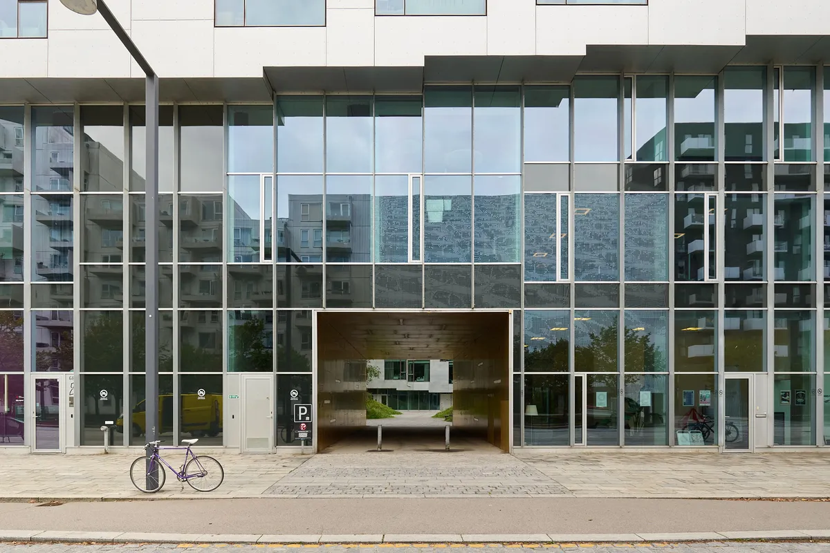 Moderne glasbygning med stor garageindkørsel og parkeringsplads foran. Glasvinduer, stålsøjler og parkerede cykler i forgrunden. Tidligere kontorlokaler i baggrunden.