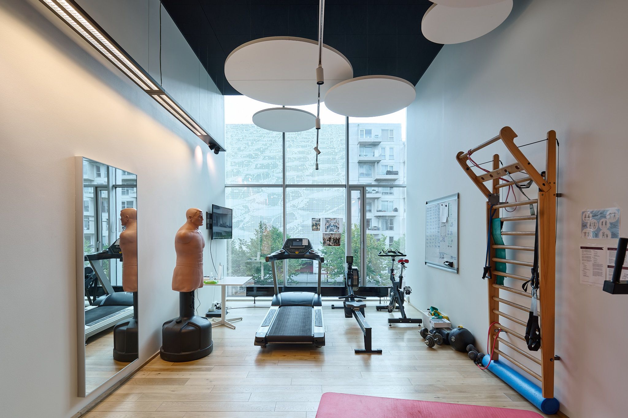 Fitnesscenter med trægulv, tre løbebånd og en træningstrappe. Træstol, træningstræ, ruder og søjler. I spejlet reflekteres en træningstræ og en mandstors dukke.