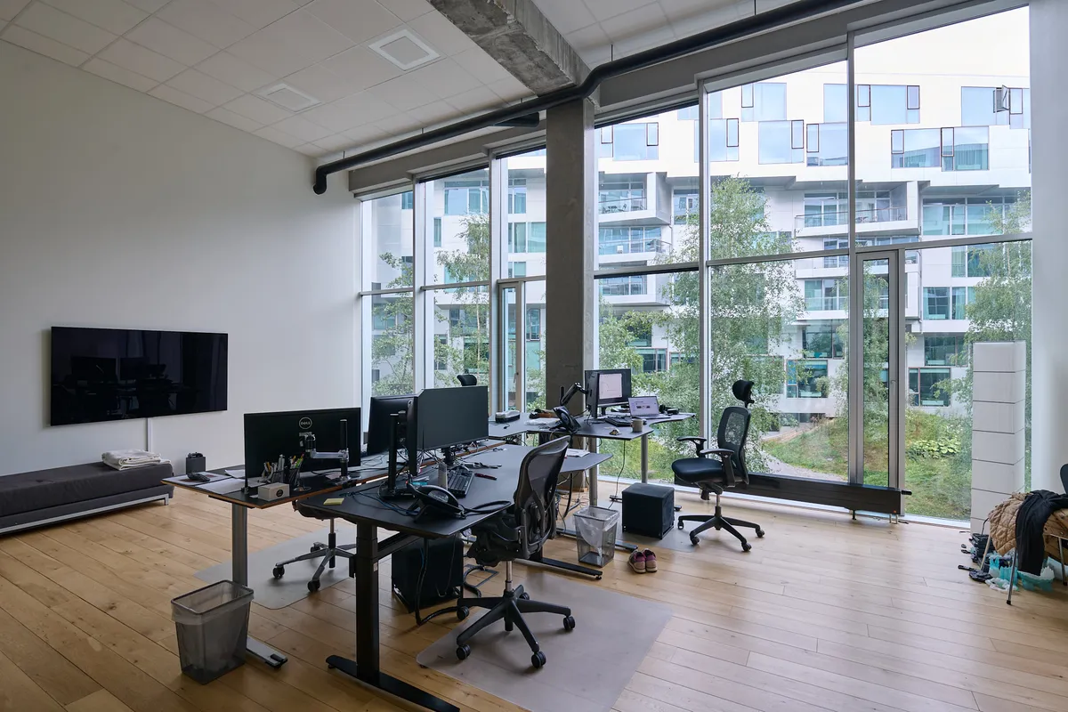 Kontor med flere skriveborde og monitorer, store vinduer og en udsigt til grønne områder og bygninger