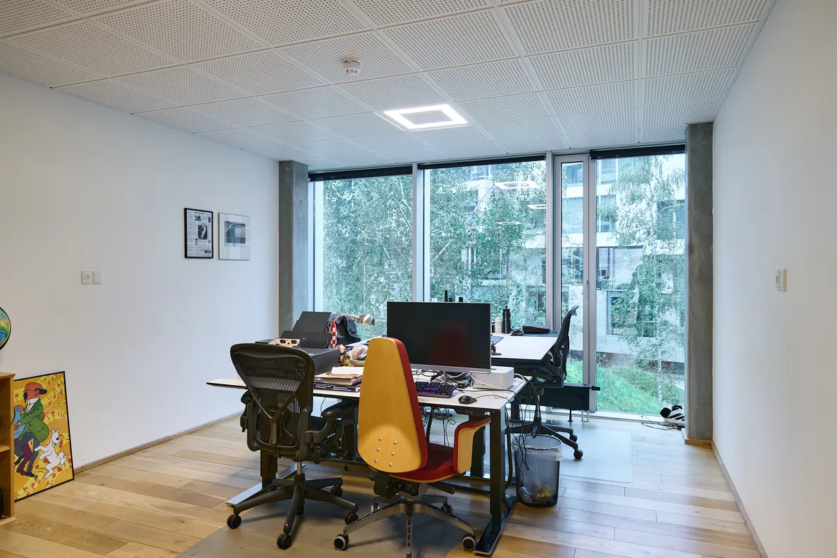 Kontor med højt gulv, hvide vægge og lyse lofter. Bord med computer og flere sæder. Vinduer med udsigt til grønne træer.