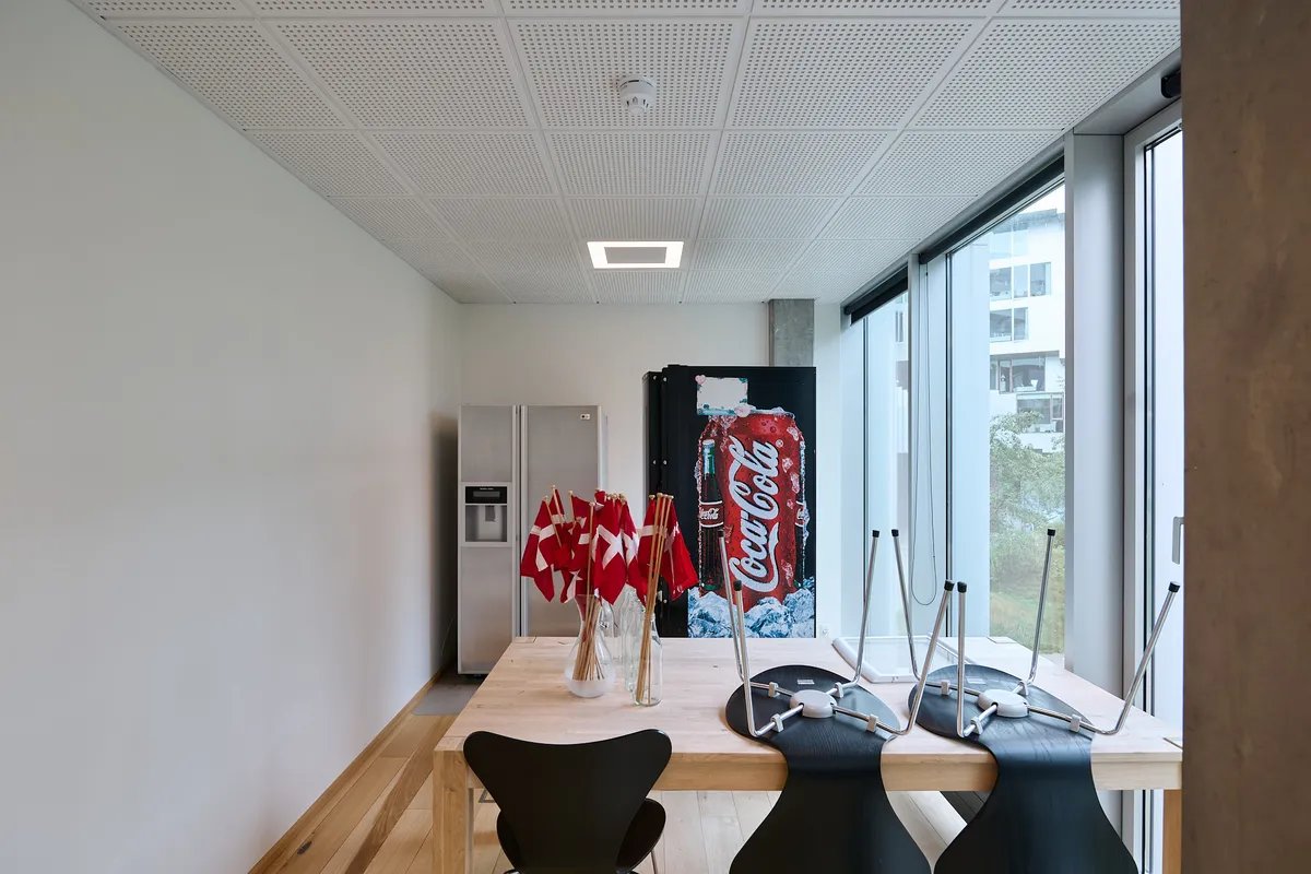 Et moderne kontor med massivt bord og flag i vase. Væggene er hvide, og der er et køleskab og sodavandsautomat i rummet