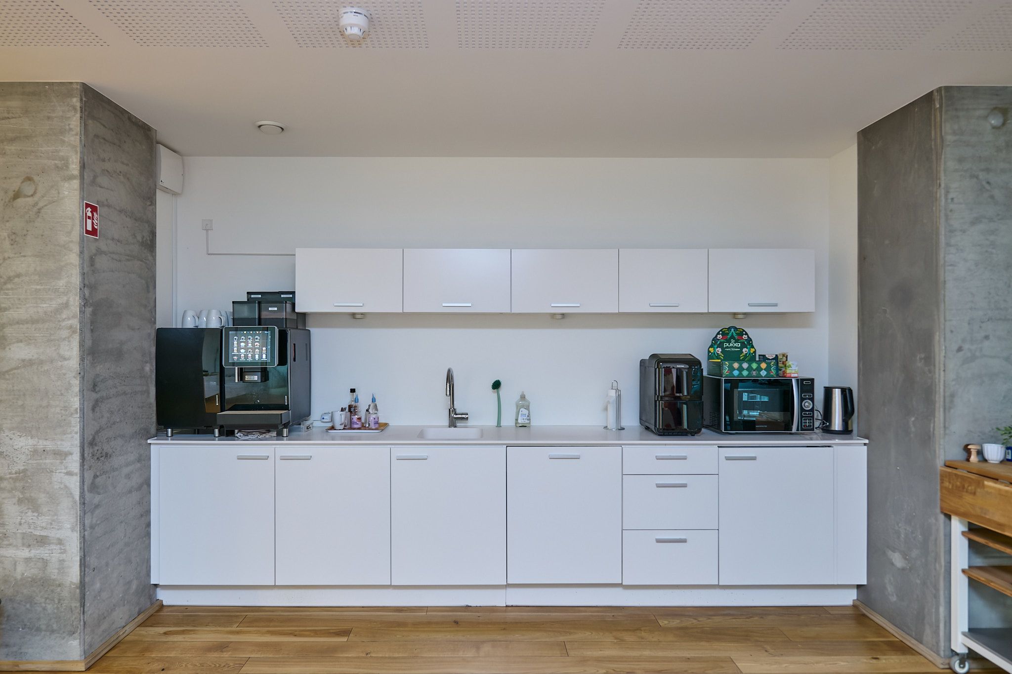 Køkken med hvide skabe og hårdt trægulv. Væggene er hvide og møblerne er moderne og minimalistiske. Der er en kaffemaskine og en mikroovn på højt skab