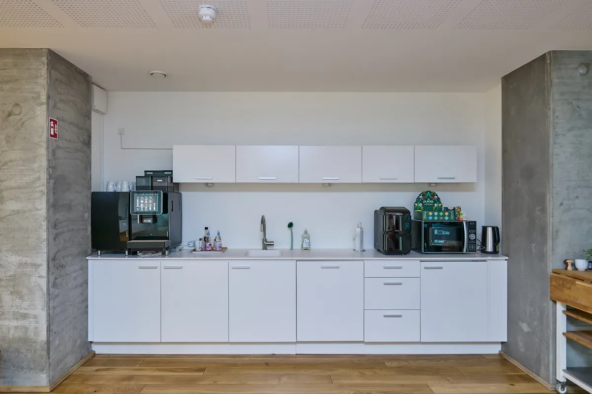 Køkken med hvide skabe og hårdt trægulv. Væggene er hvide og møblerne er moderne og minimalistiske. Der er en kaffemaskine og en mikroovn på højt skab