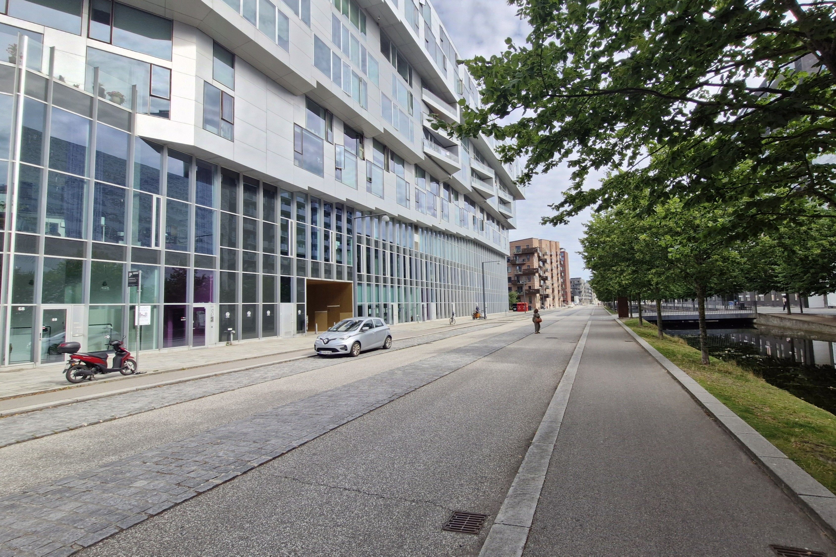 Moderne kontorbygning med glasfacade. Parkvej med grønne træer og enkelte gående. Parkerede scooter og bil. Bygning med høje vinduer og skyggede områder