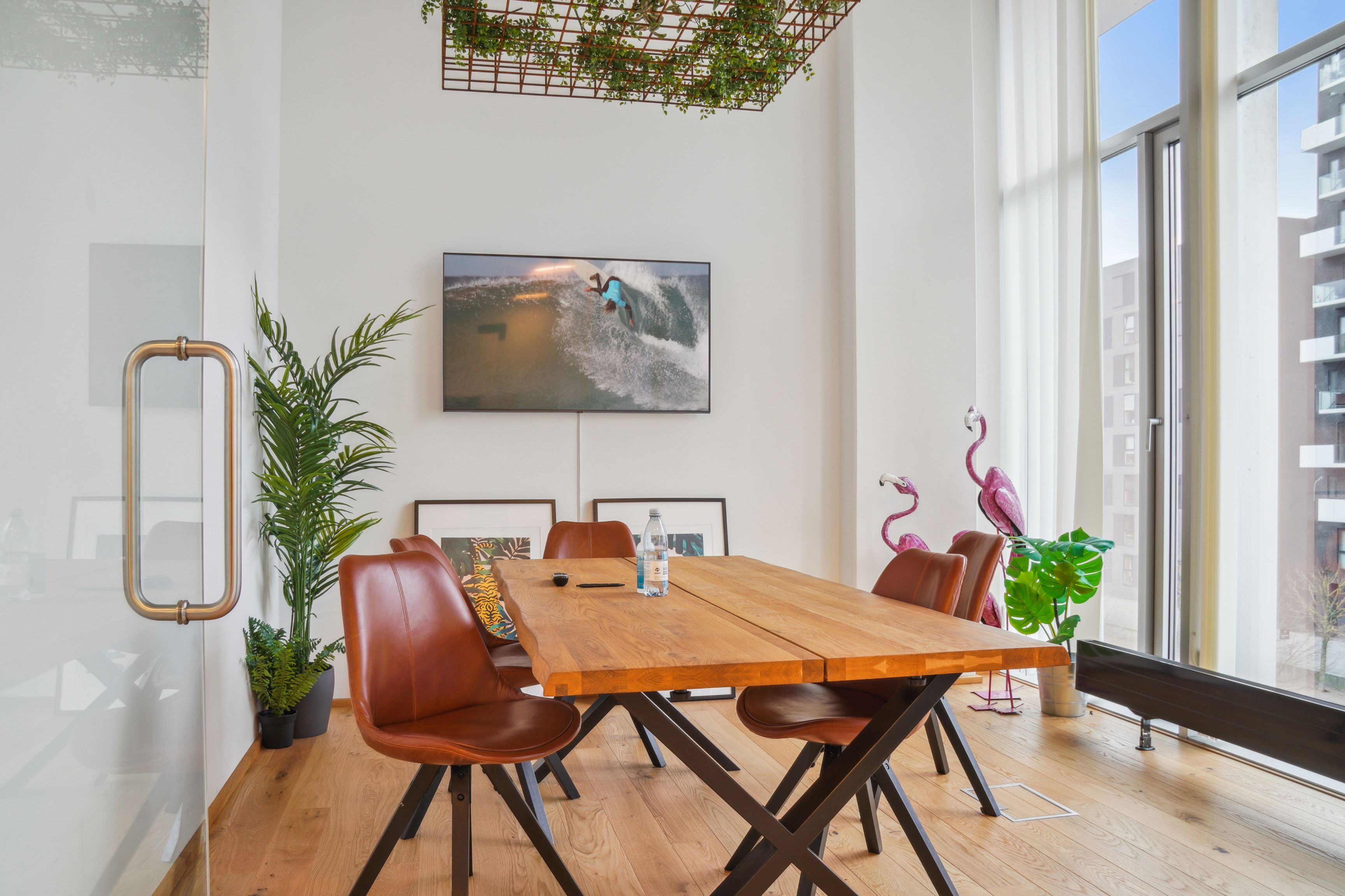 Kontor med træbord, læderstole og plante. Store ruder med udsigt til bygninger og plante. Vægmonteret TV-skærm med surfer på bølger.